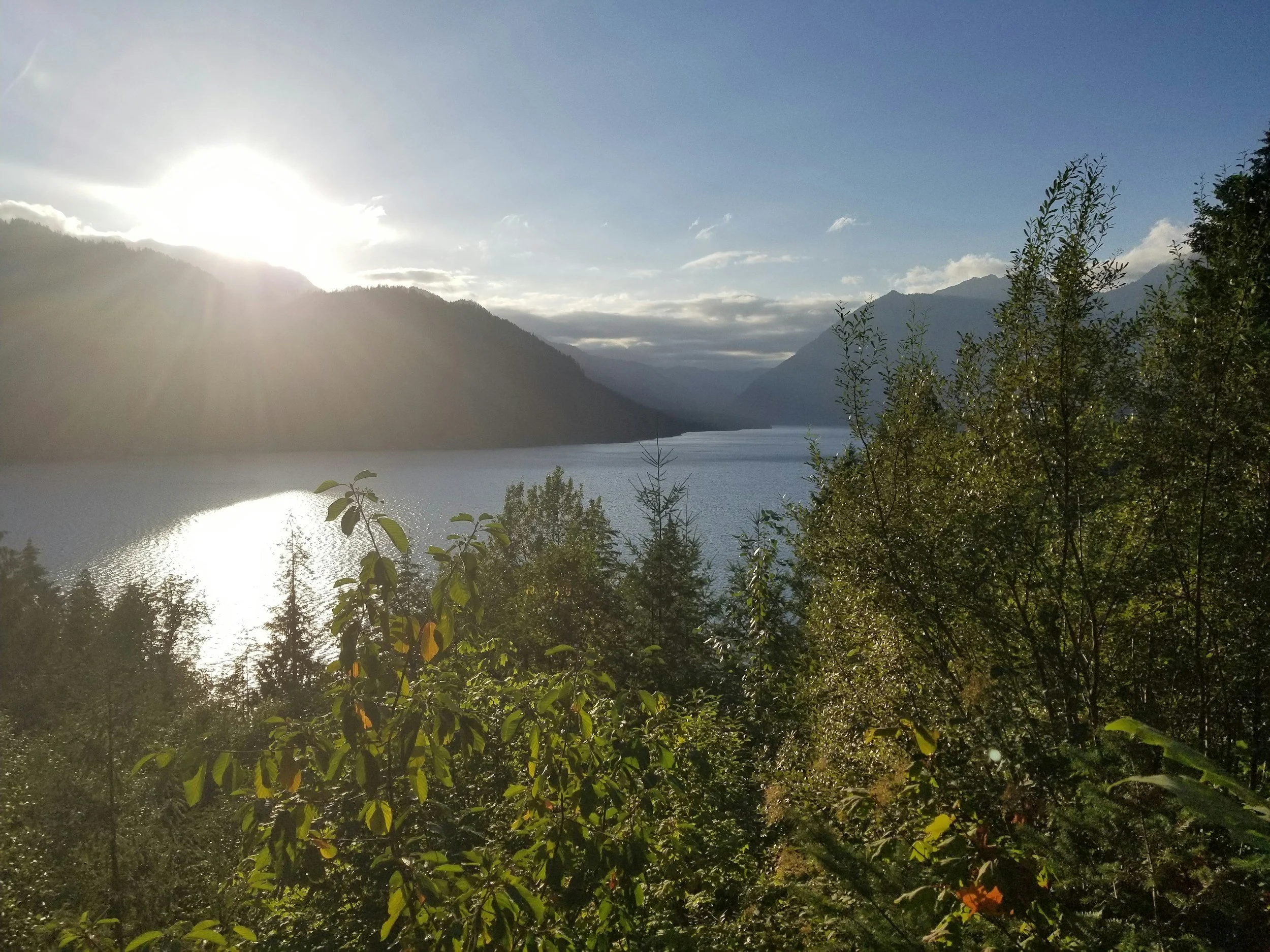 Sunrise or sunset over a mountain lake with trees in the foreground and mountains in the background.