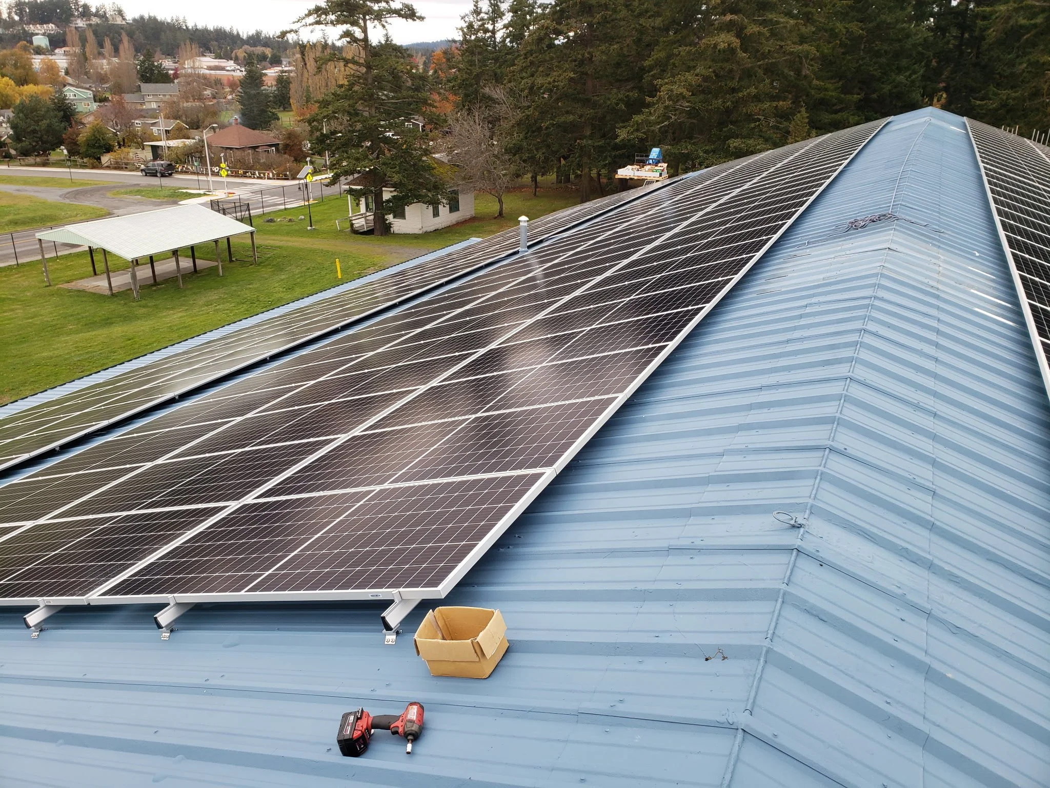 On the roof of a building, installed solar panels are visible, along with tools, a cordless drill, and a cardboard box, indicating ongoing installation work.