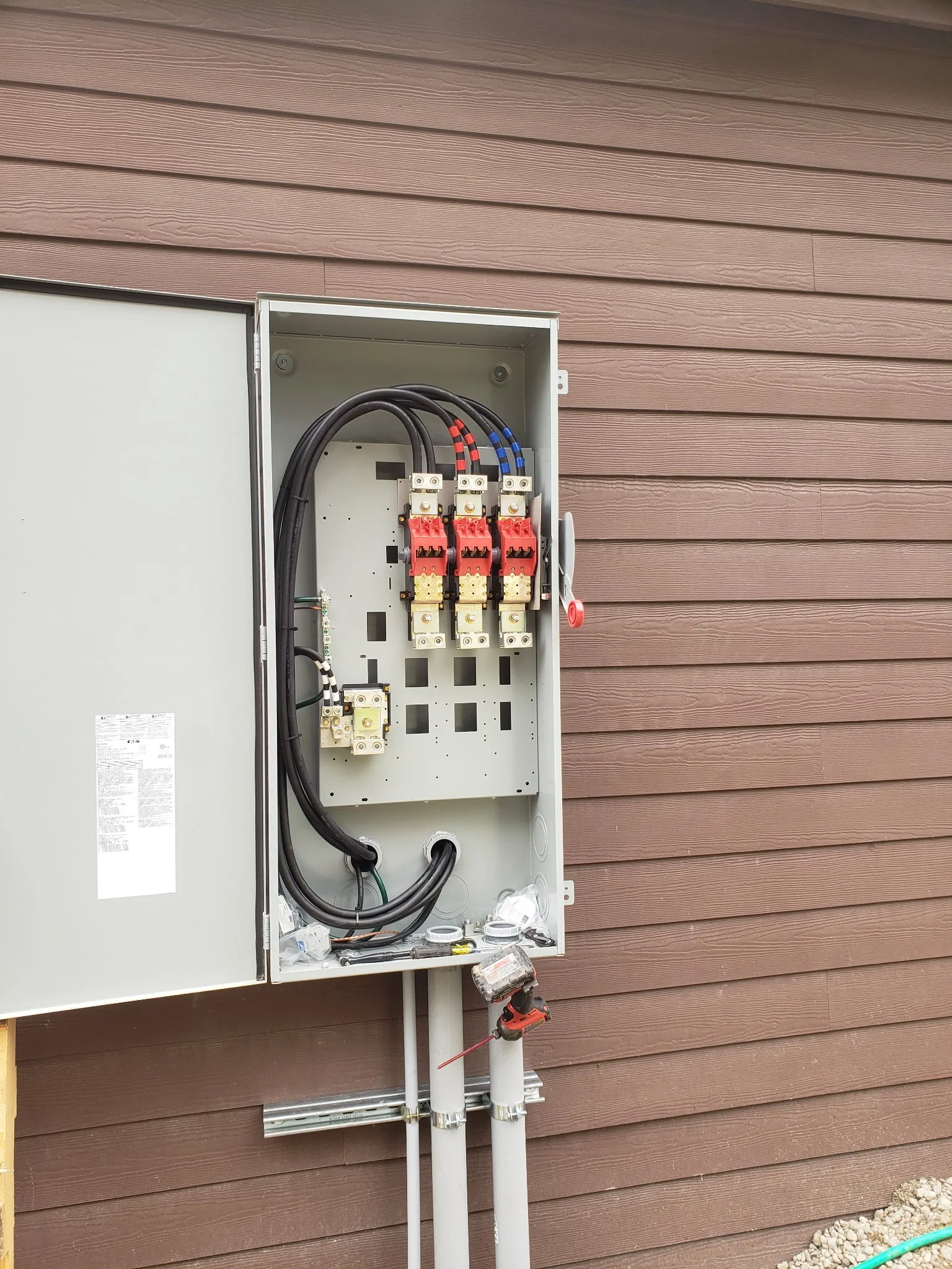 Outdoor electrical panel with wiring and connections, mounted on a brown wooden wall, with tools and materials at the bottom.
