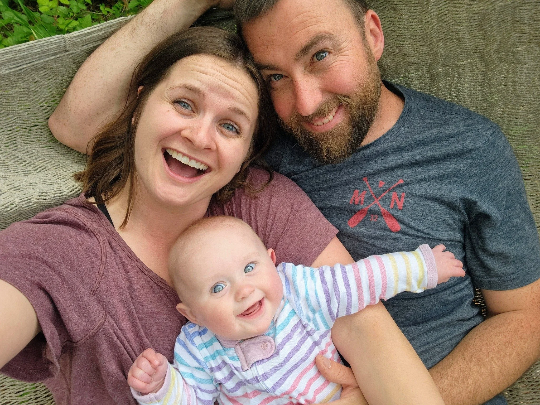 A happy family of four, including a woman, a man, and two young children, lying together outdoors on a blanket, smiling at the camera.