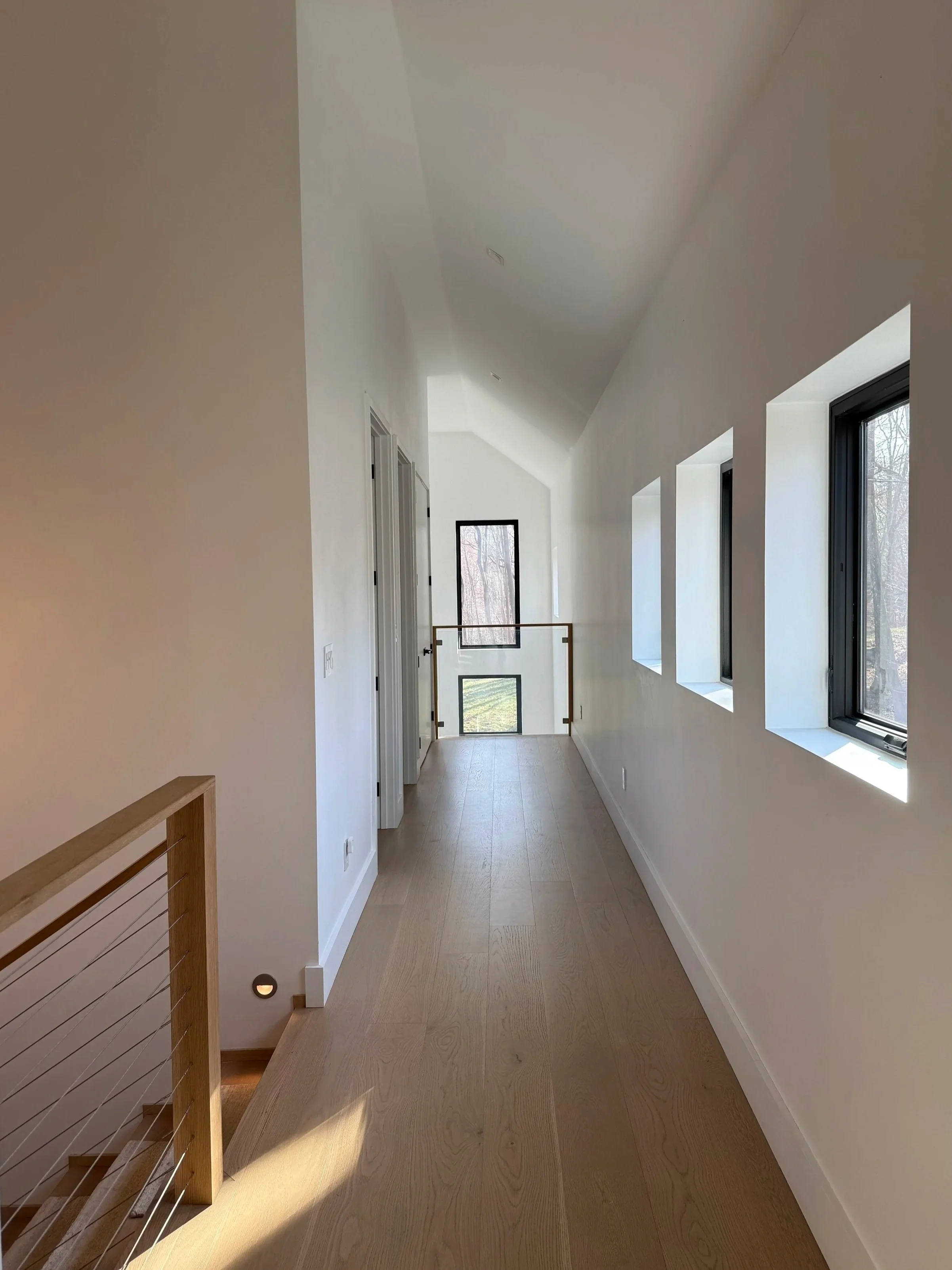 A narrow hallway with white walls, three rectangular windows on the right, and a staircase with wooden railing leading down on the left. The end of the hallway features a door or window with a view of trees outside.