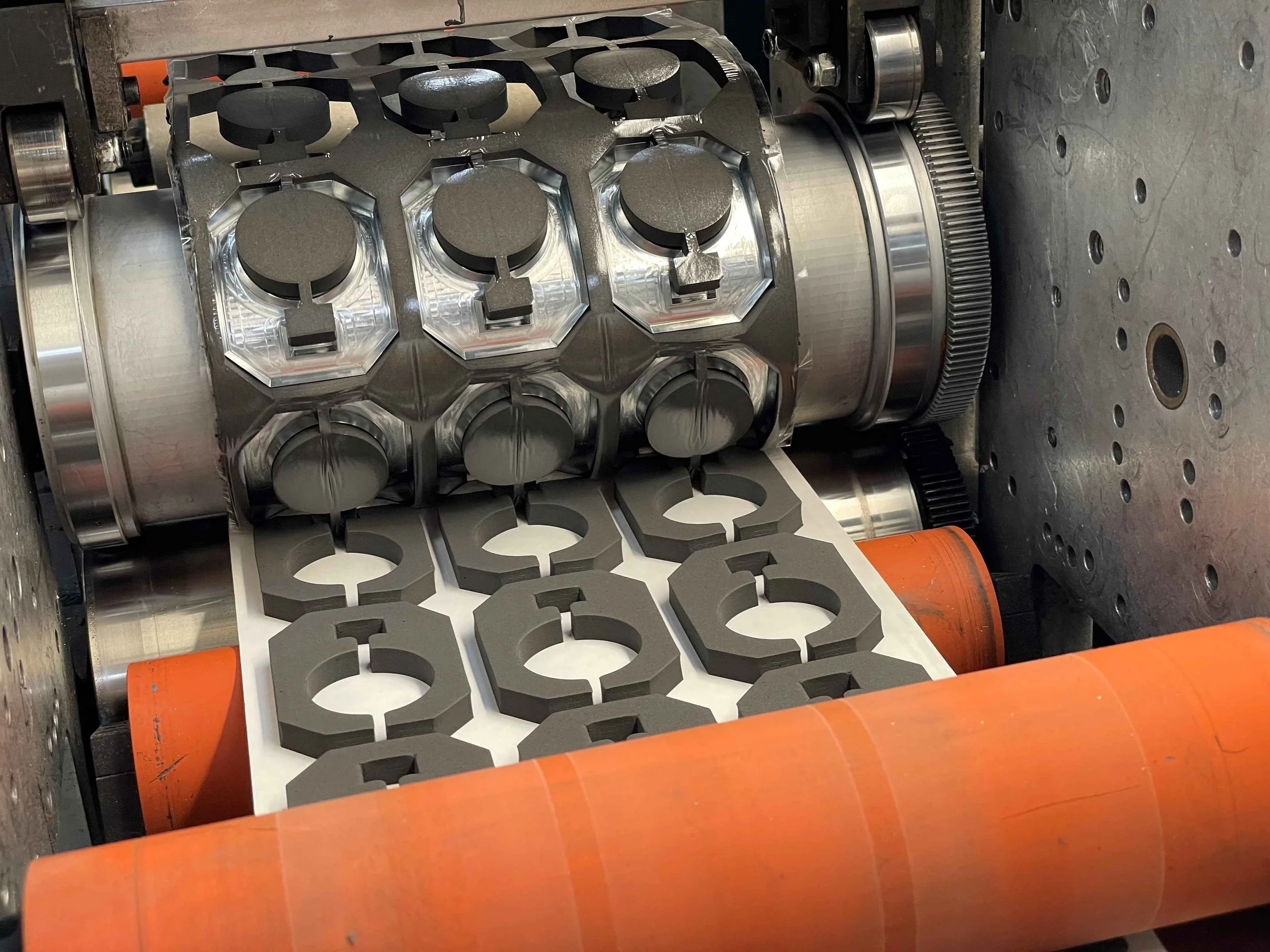 Close-up view of a manufacturing process involving a machine stamping or cutting hexagonal and circular rubber or foam gaskets from sheets of material. The process includes rollers and metal components.