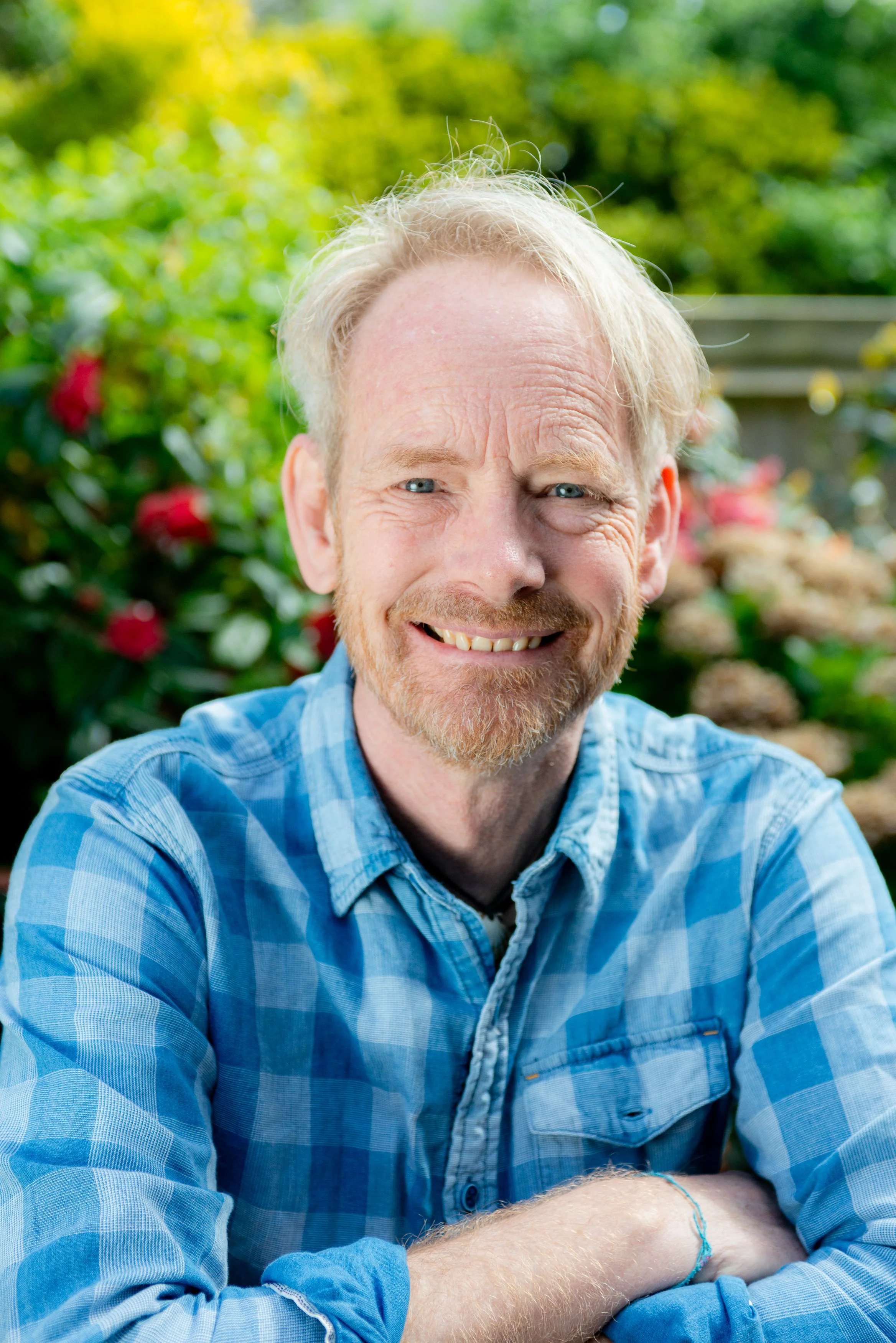 A smiling middle-aged man with blonde hair and a beard, wearing a blue plaid shirt, sitting outdoors with a blurred green garden background.