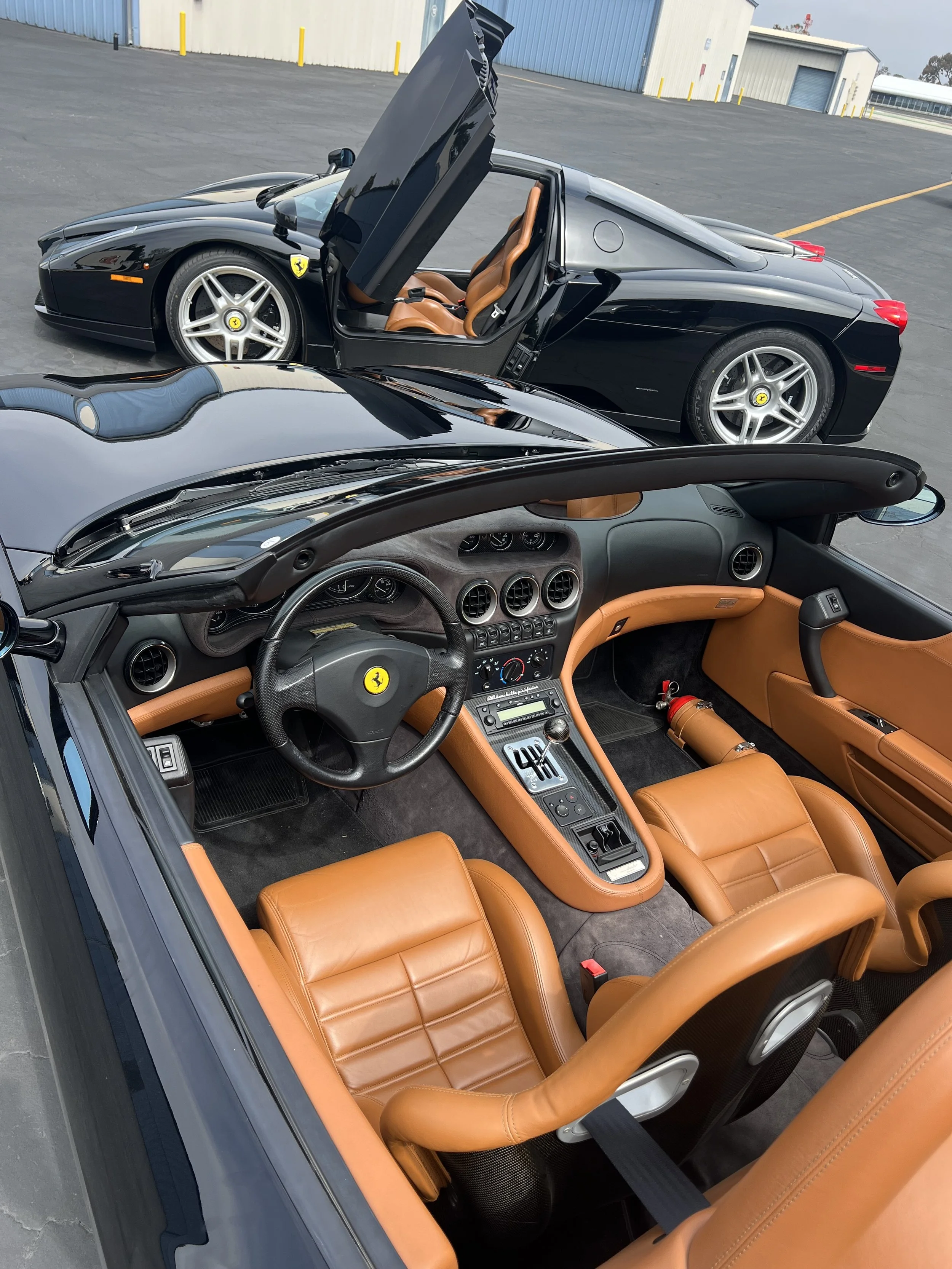 Interior of a black Ferrari with tan leather seats and dashboard, visible with a similar black Ferrari in the background with its driver-side door open, parked in an open lot next to industrial buildings.