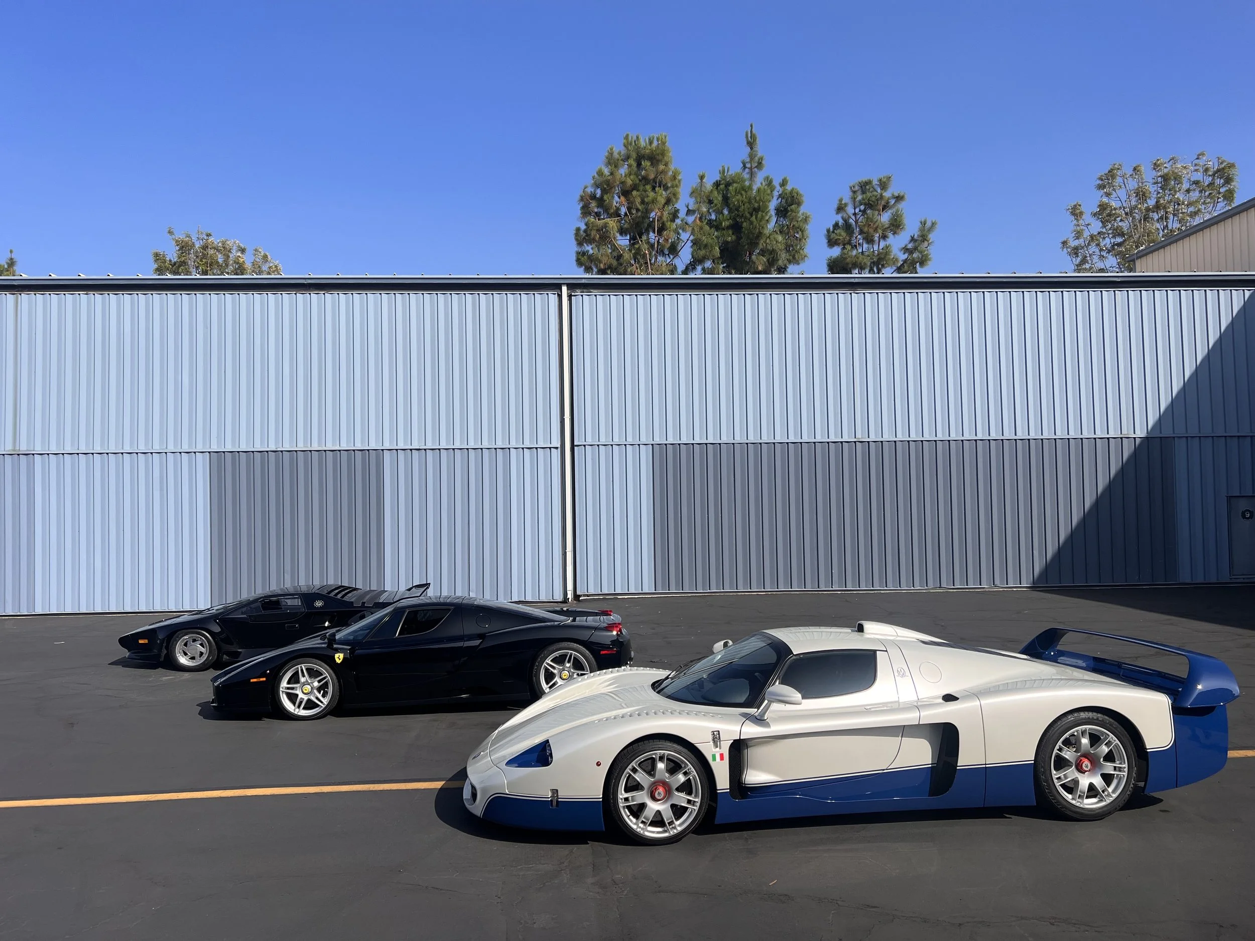 Three sports cars parked on a paved lot in front of a large blue metal building under a clear blue sky.