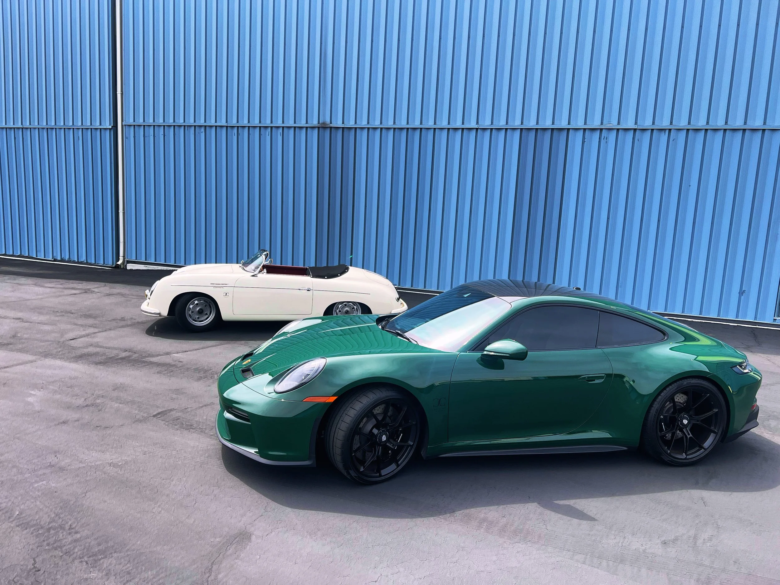 A green sports car and a white vintage convertible parked in front of a blue corrugated metal wall.