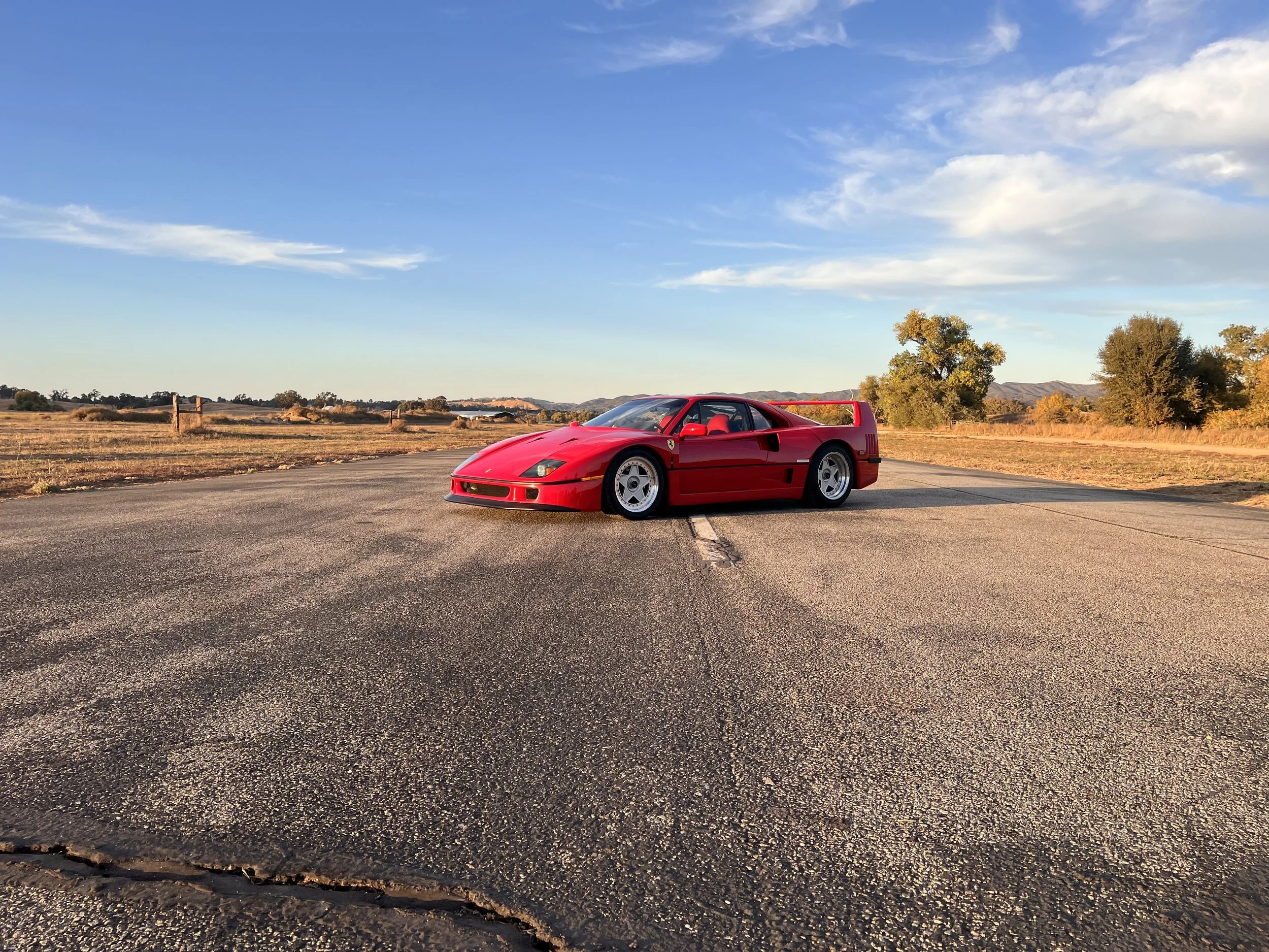 Red Ferrari F40 sports car parked on an empty asphalt road in a rural landscape with trees and hills in the background during sunset.