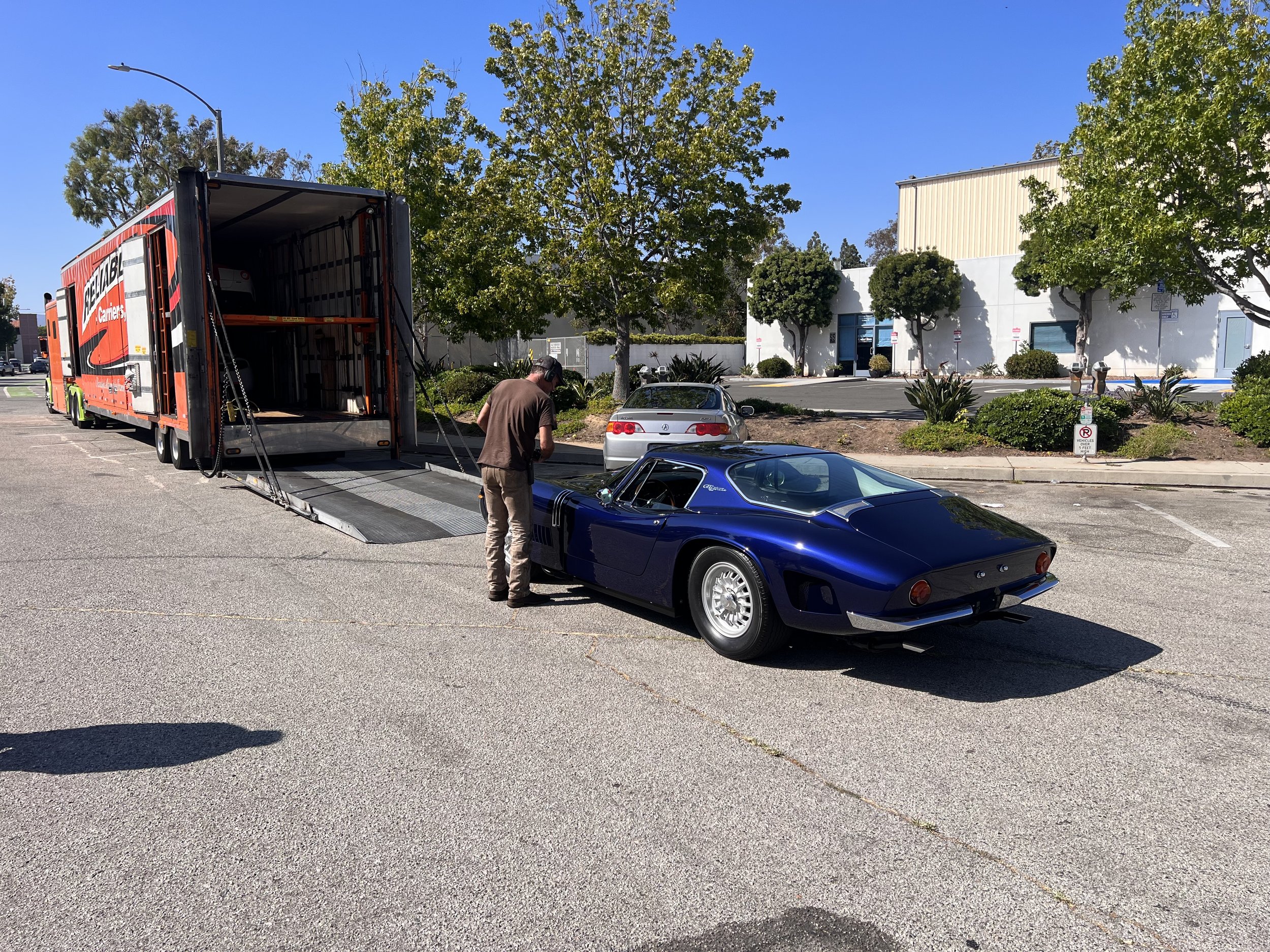 A person standing beside a classic blue sports car in a parking lot, loading it into a large transportation truck with an open ramp.