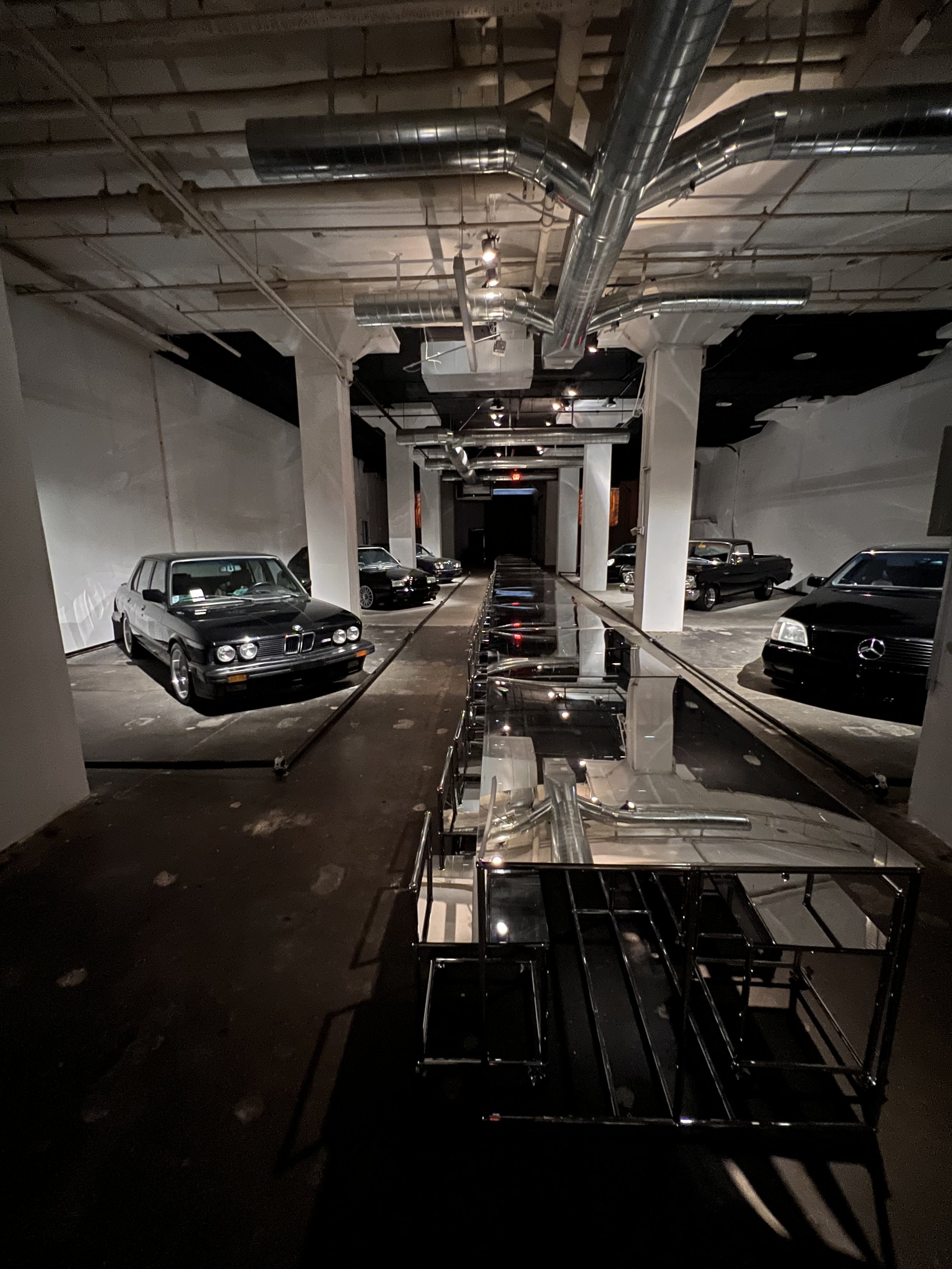 An indoor car showroom with several classic cars on display, including BMW and Mercedes-Benz models, under industrial-style ceiling with exposed ducts.