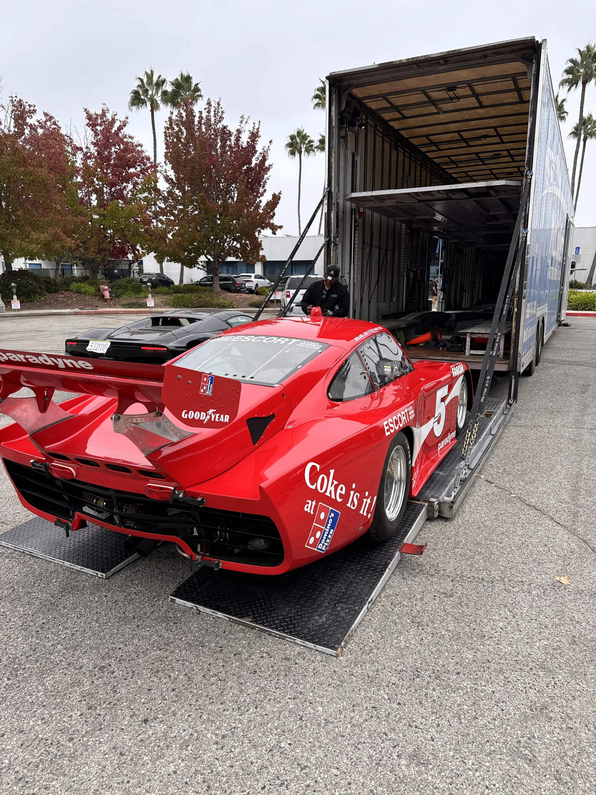 A red race car with sponsorship logos, including Coke and Goodyear, positioned on a black loading ramp. A person in black attire is standing by the car, with a large open car carrier trailer behind it, ready for transport. The setting appears to be o