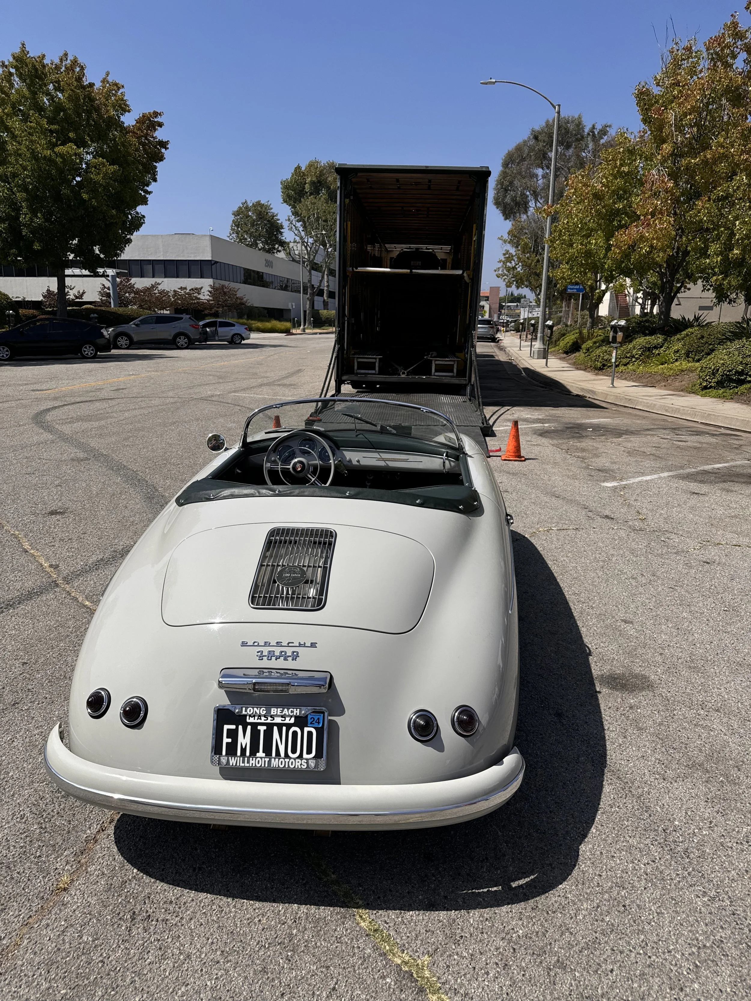 A vintage white Porsche 356 convertible parked in an outdoor lot, connected to a large transport truck.