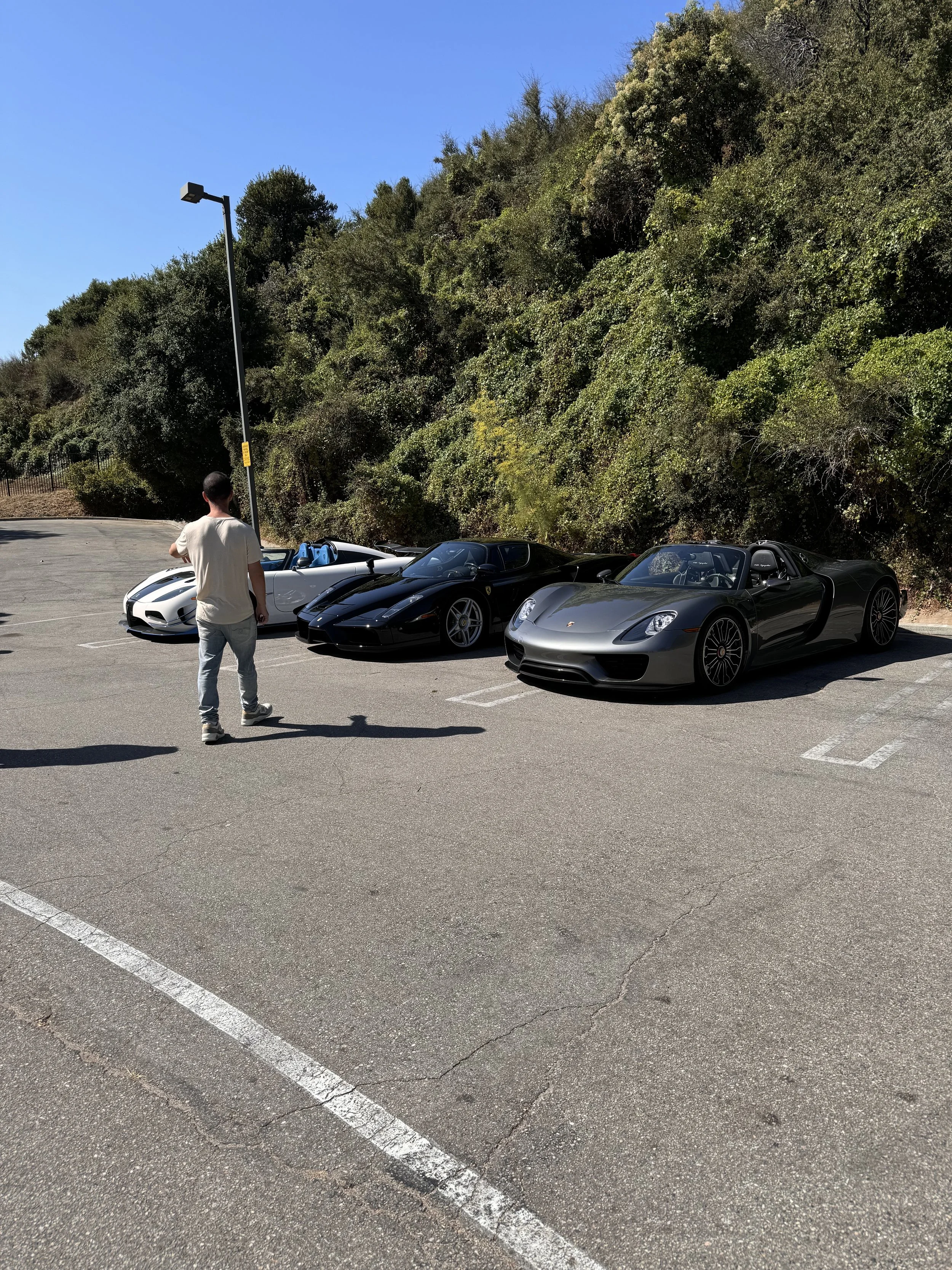Three luxury sports cars parked in a parking lot next to a hillside covered in greenery, with a man standing nearby and looking at the cars.