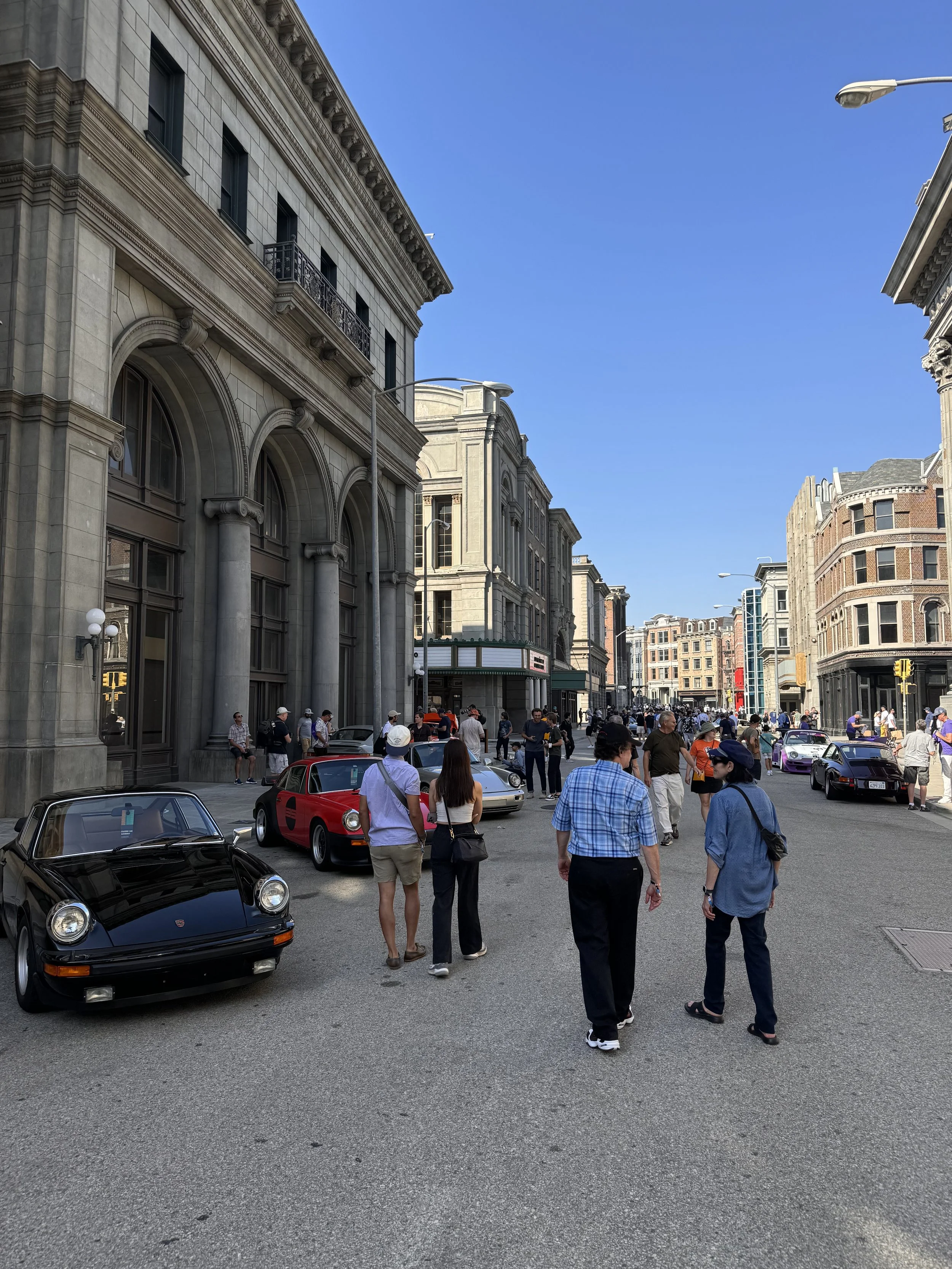 A city street filled with people and parked classic cars, including a black Porsche, on a sunny day with large historic buildings.