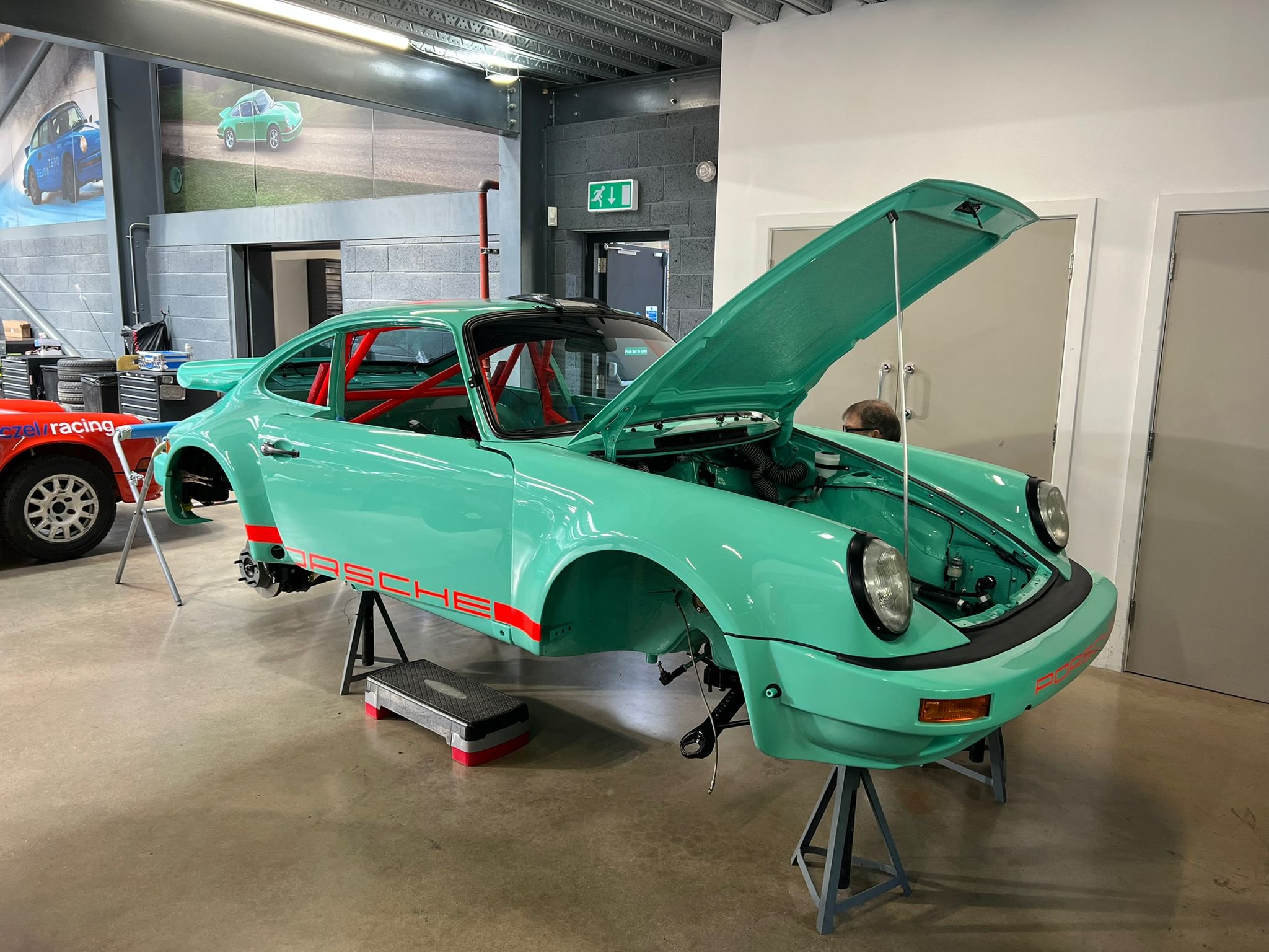 A vintage Porsche race car in a mint green color, with its hood open and wheels removed, inside a workshop or garage. The car has red accents and the Porsche logo on the side.