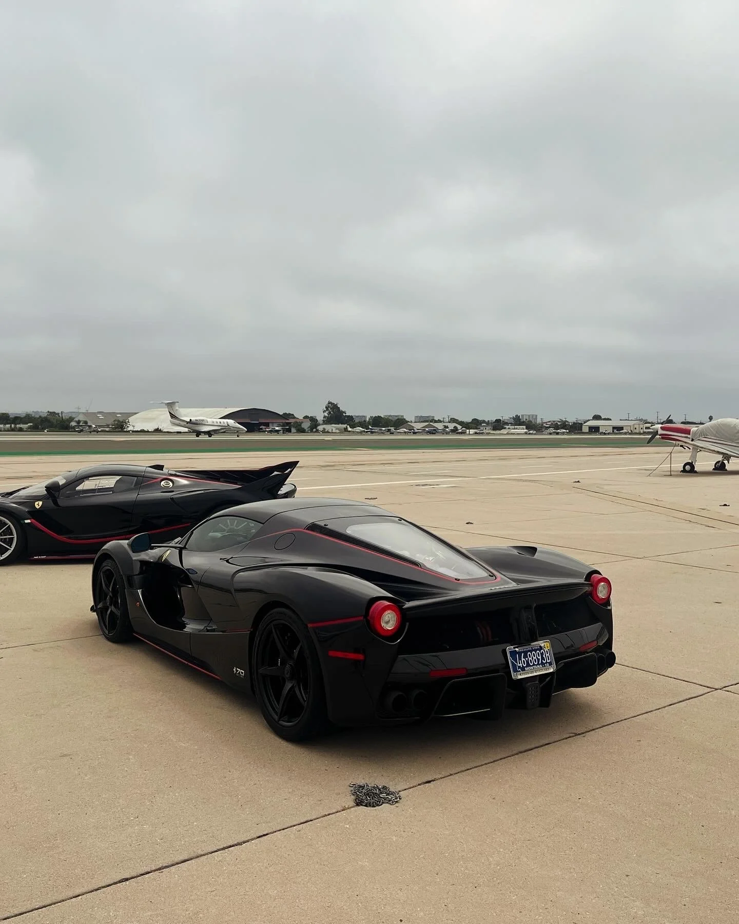 Two black sports cars parked on an airport tarmac with private jets and aircraft hangars in the background under grey, overcast skies.
