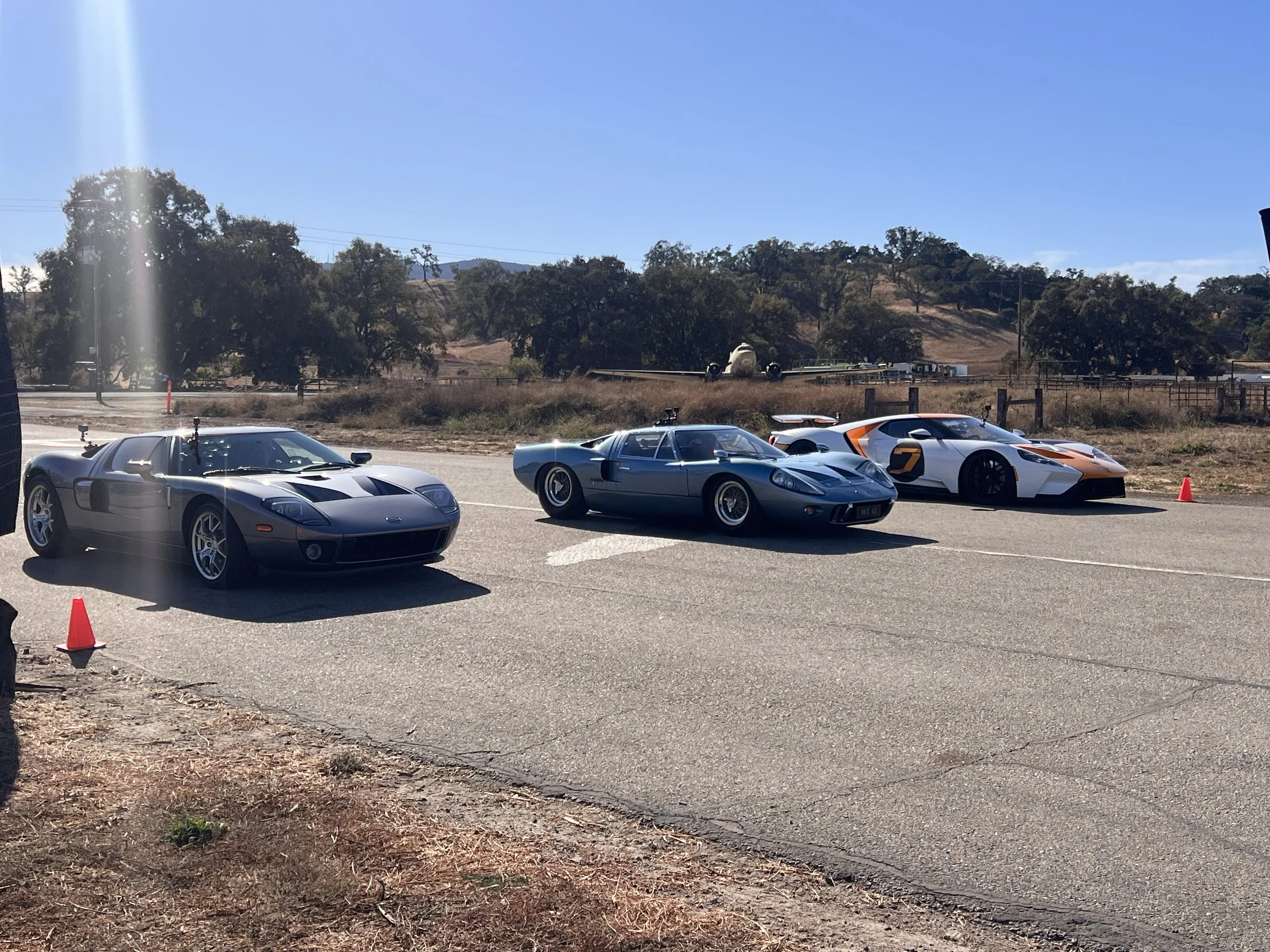 Three sports cars parked on the side of a road with orange cones behind them, with a scenic background of trees, hills, and a plane on the runway under a clear blue sky.