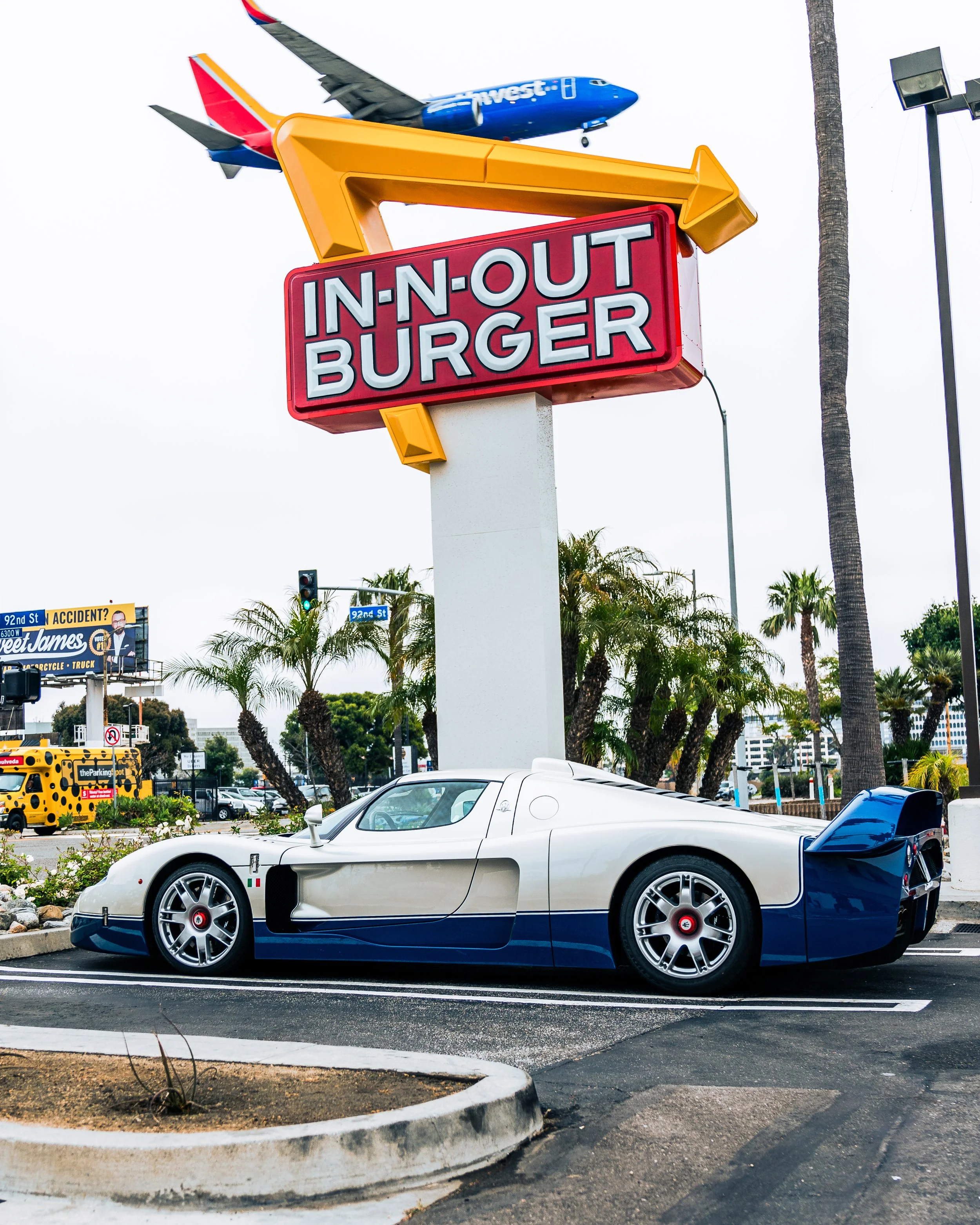 A white and blue racing car parked in front of an In-N-Out Burger sign with palm trees and a plane flying overhead in the background.