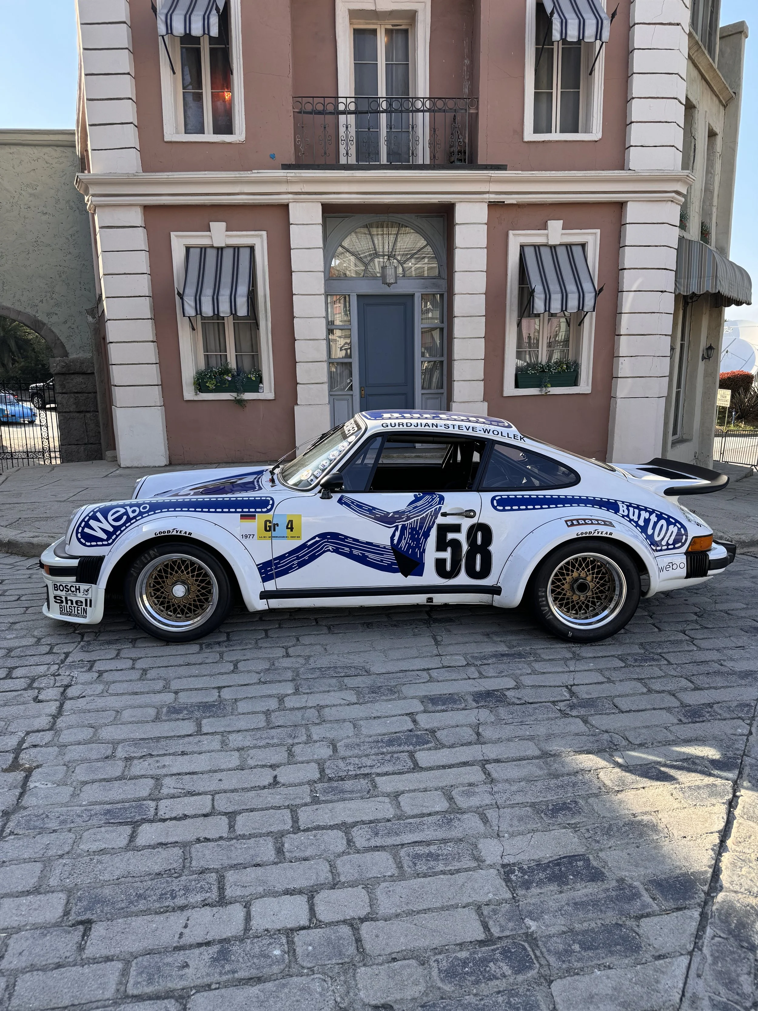 A classic Porsche race car painted in white with blue graphics and sponsor logos, parked on a cobblestone street in front of a pink and beige townhouse with striped awnings and a blue door.