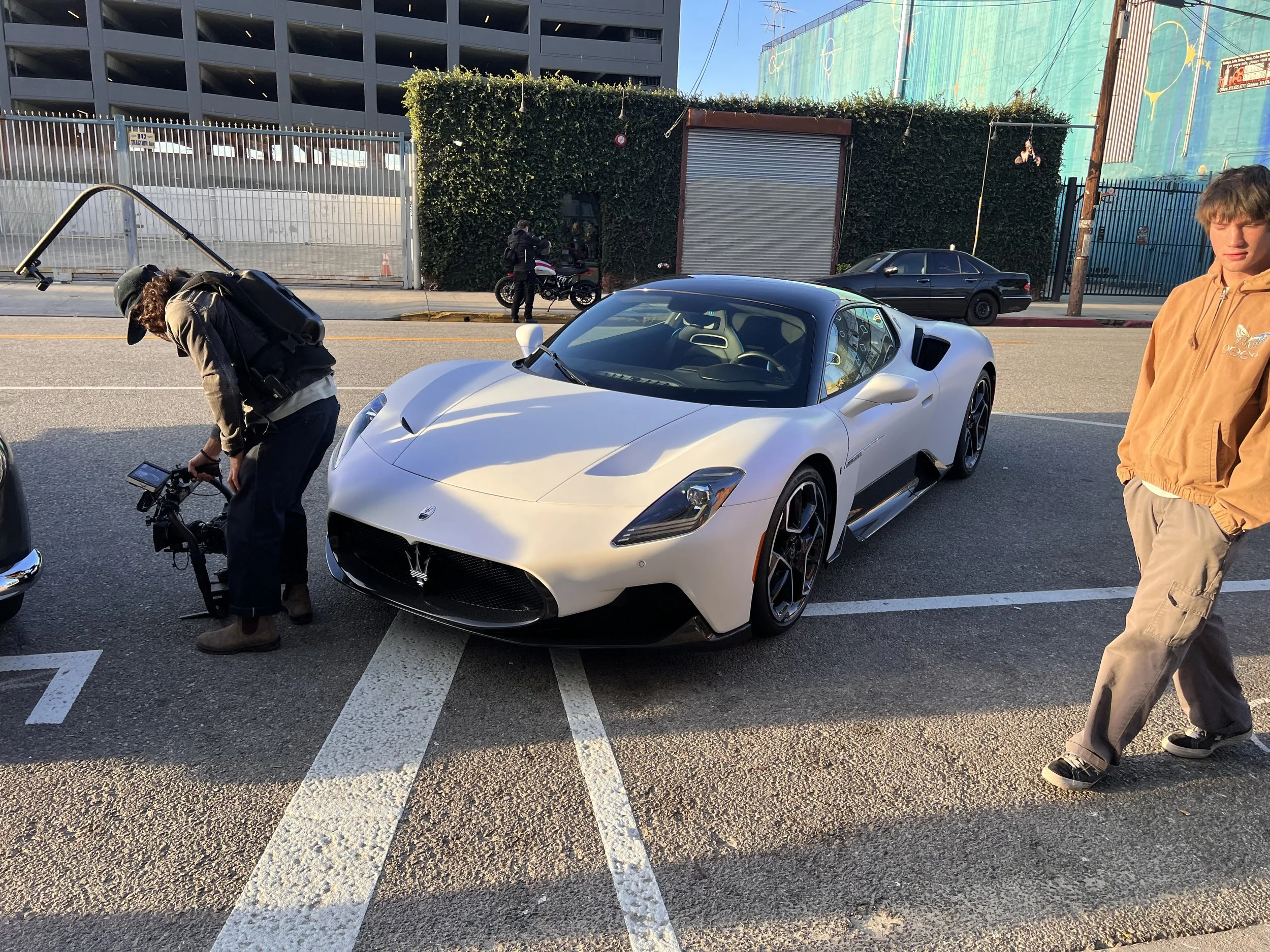 A white Maserati sports car parked on the street with two men nearby; one is adjusting a camera, the other is walking past; there is a scooter and another car in the background.