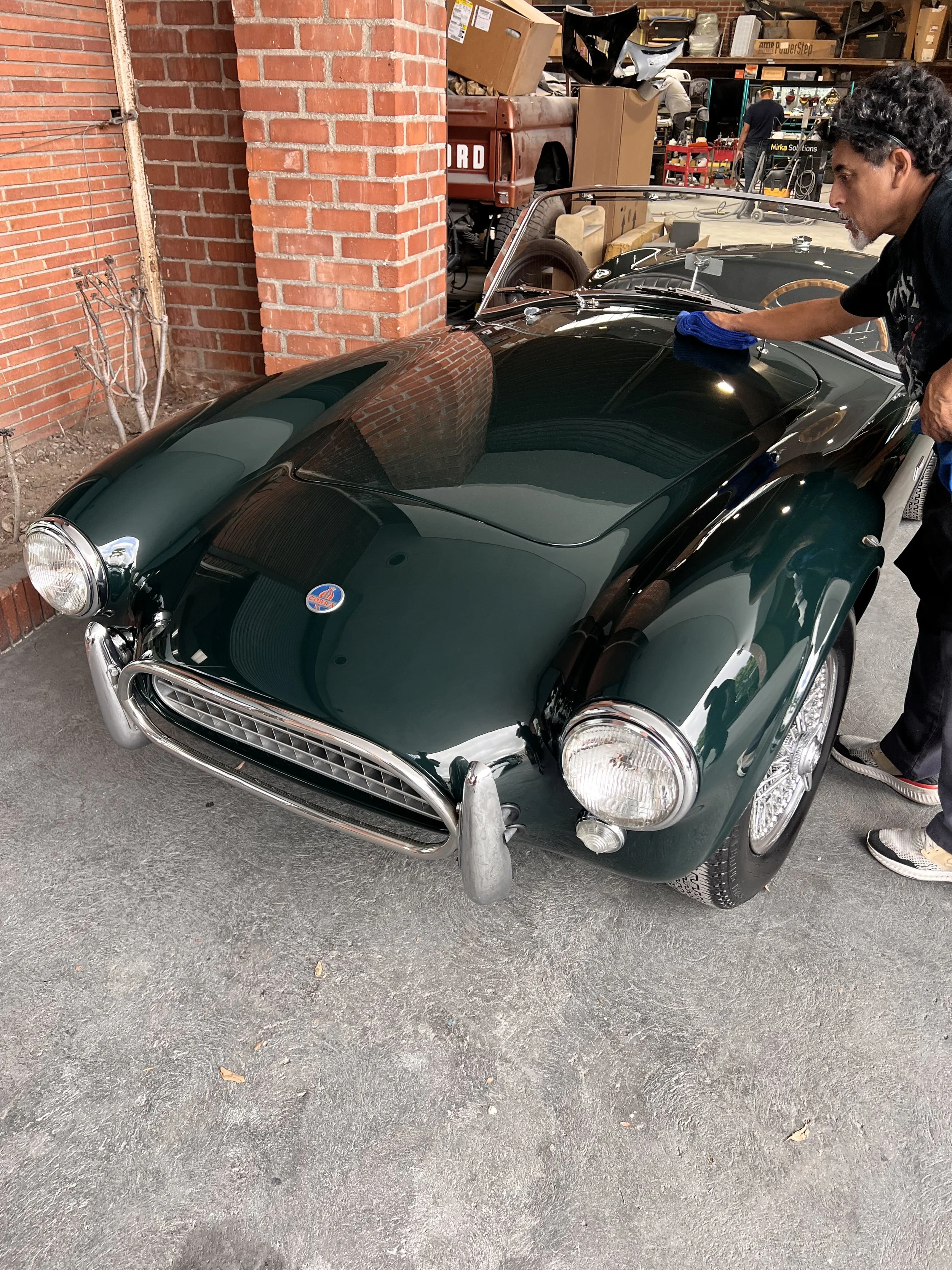 A man is polishing a vintage dark green sports car with a blue cloth inside a garage with brick walls and various boxes and tools in the background.