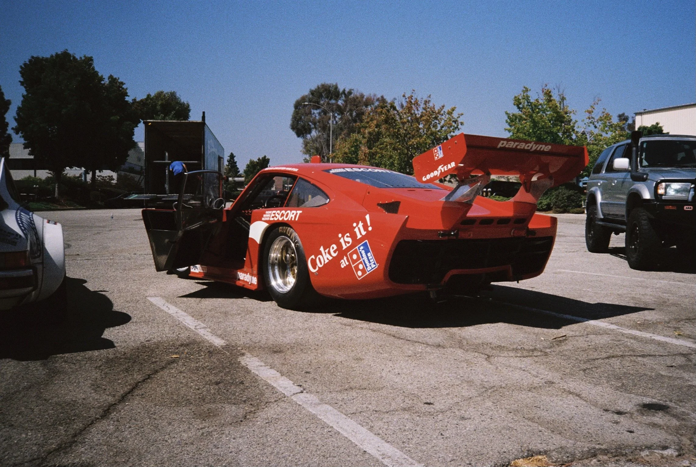 Red race car with advertisements for Coke and Domino's pizza parked in a parking lot, with other vehicles and trees in the background.