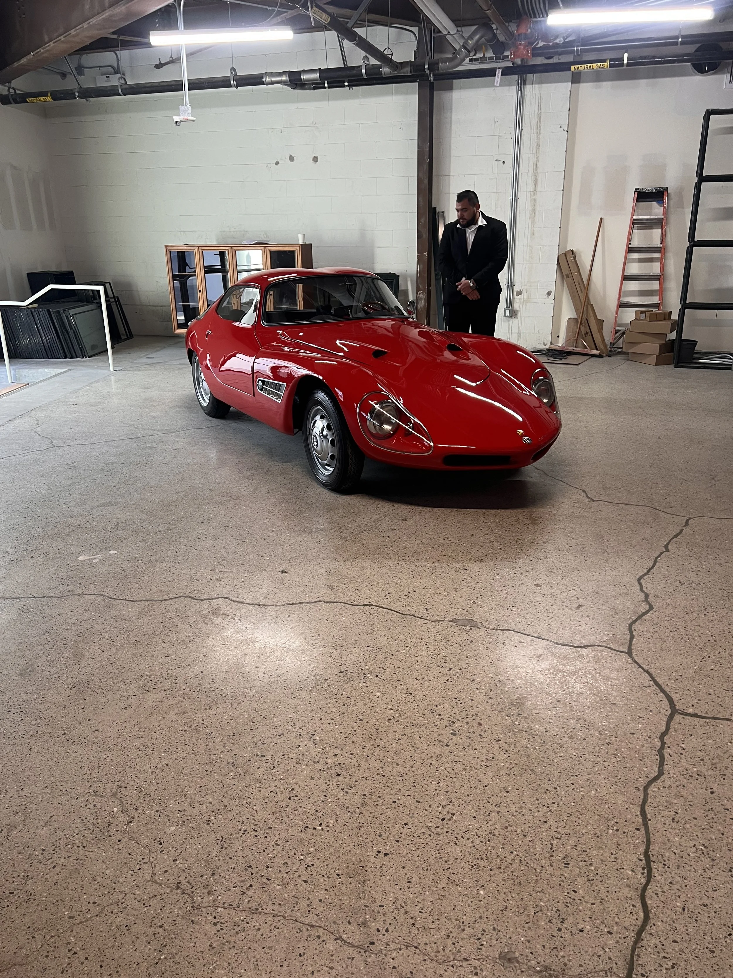 A red vintage race car inside a garage with a man in a suit standing behind it. The garage has a white brick wall, some shelves, and boxes in the background.