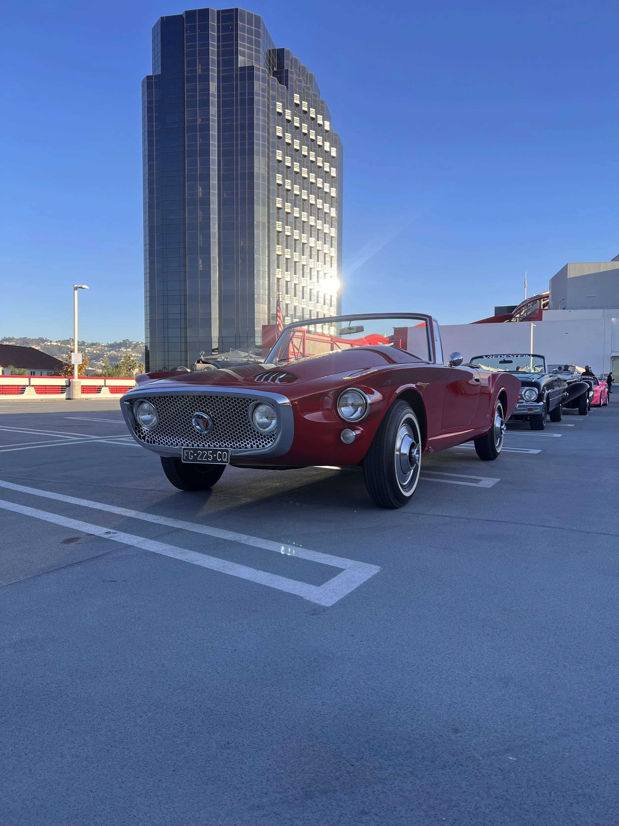 Red vintage convertible car parked in an outdoor lot with a tall modern building in the background and other cars lined up behind it.