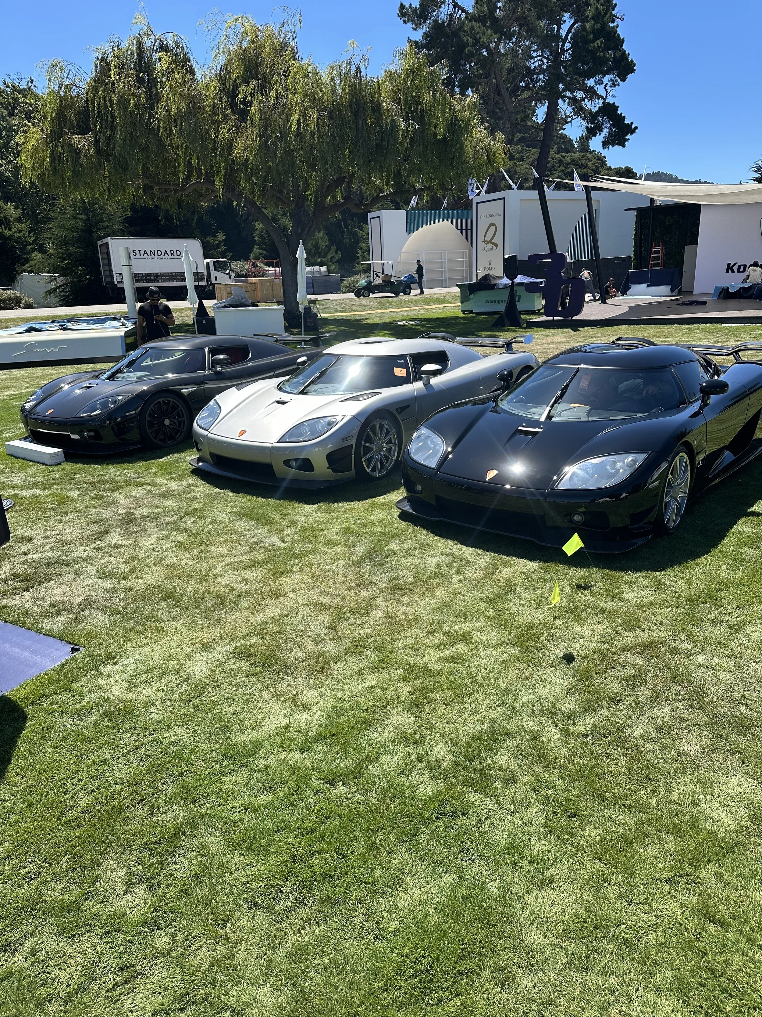 Three luxury sports cars displayed outdoors on a grassy area at an event, with tents and people in the background.