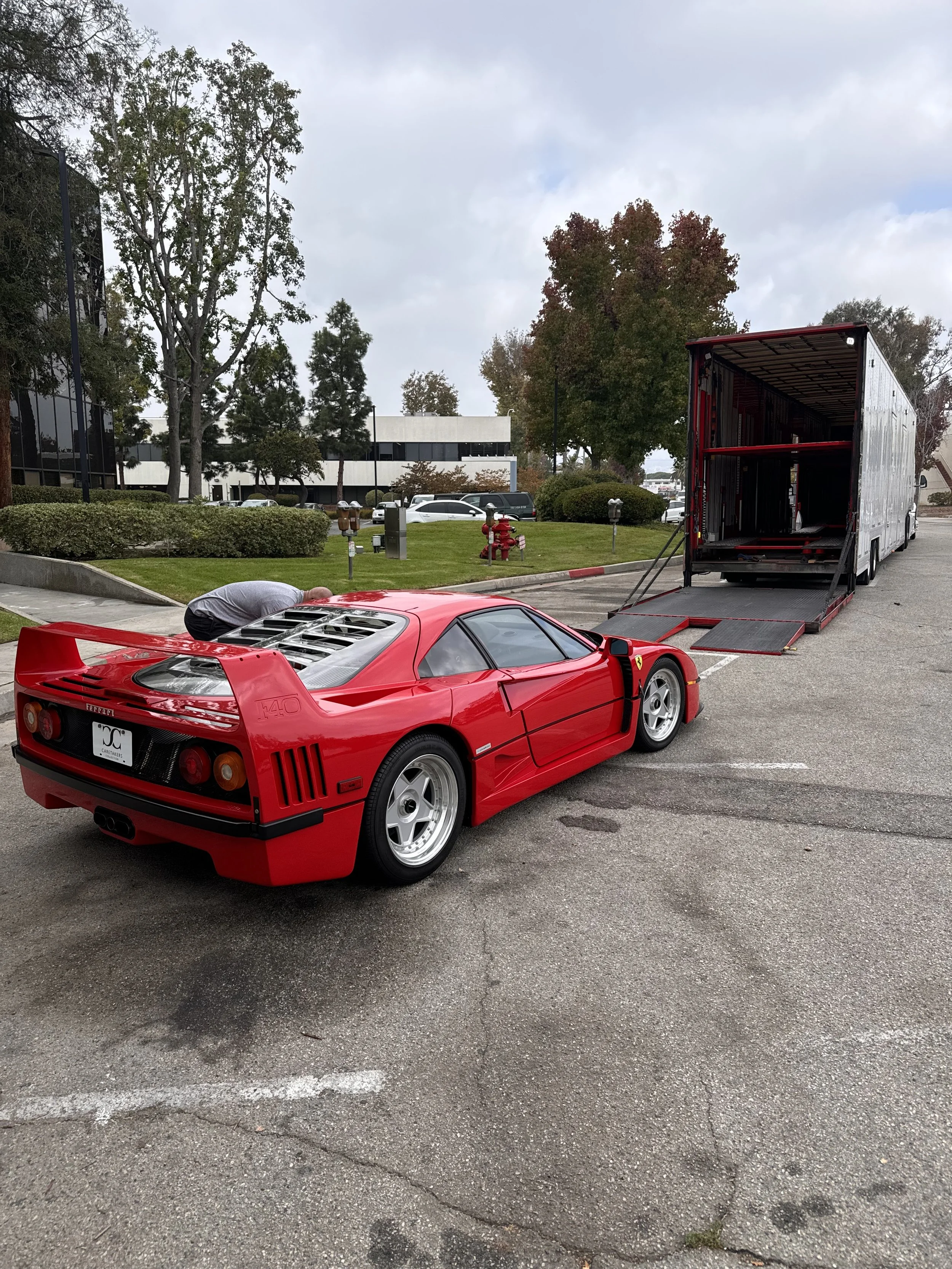 A red Ferrari F40 supercar is being loaded into a large black transporter truck in a parking lot surrounded by trees and office buildings on a cloudy day.