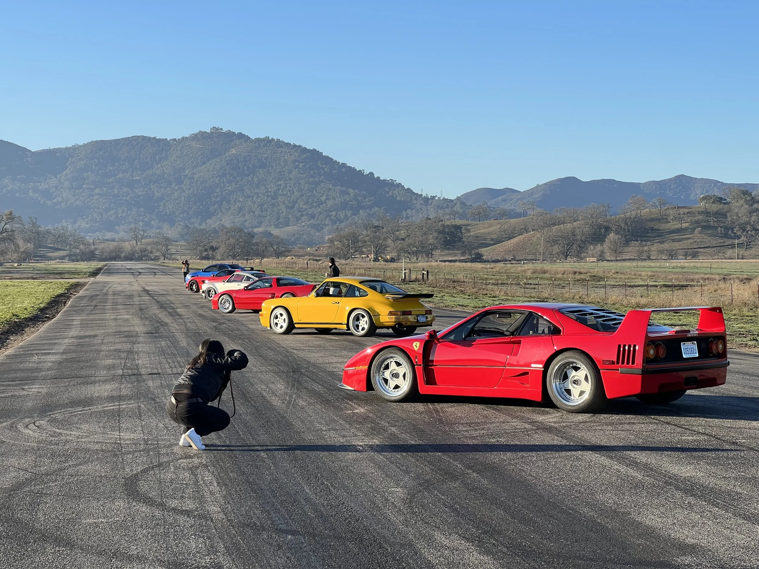 Six vintage Ferrari sports cars parked in a line on a rural road with mountains and fields in the background and two people taking photos.