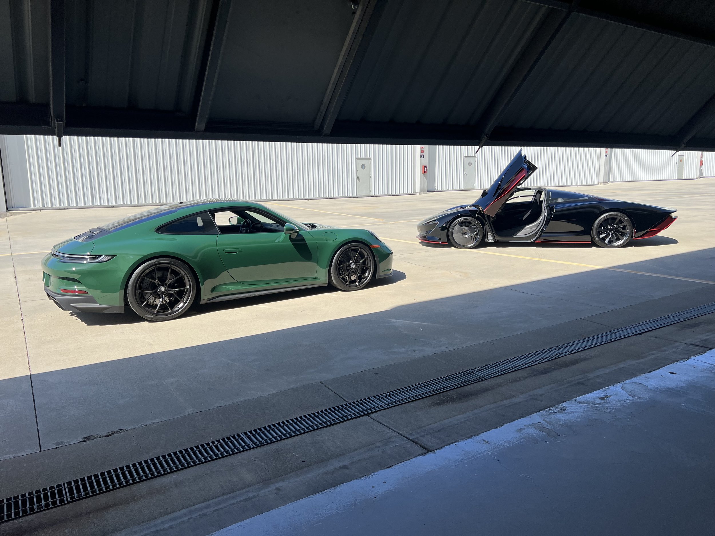 Two high-performance sports cars parked in a partially shaded parking lot, one green and one black with red accents, with the black car's gull-wing door open.