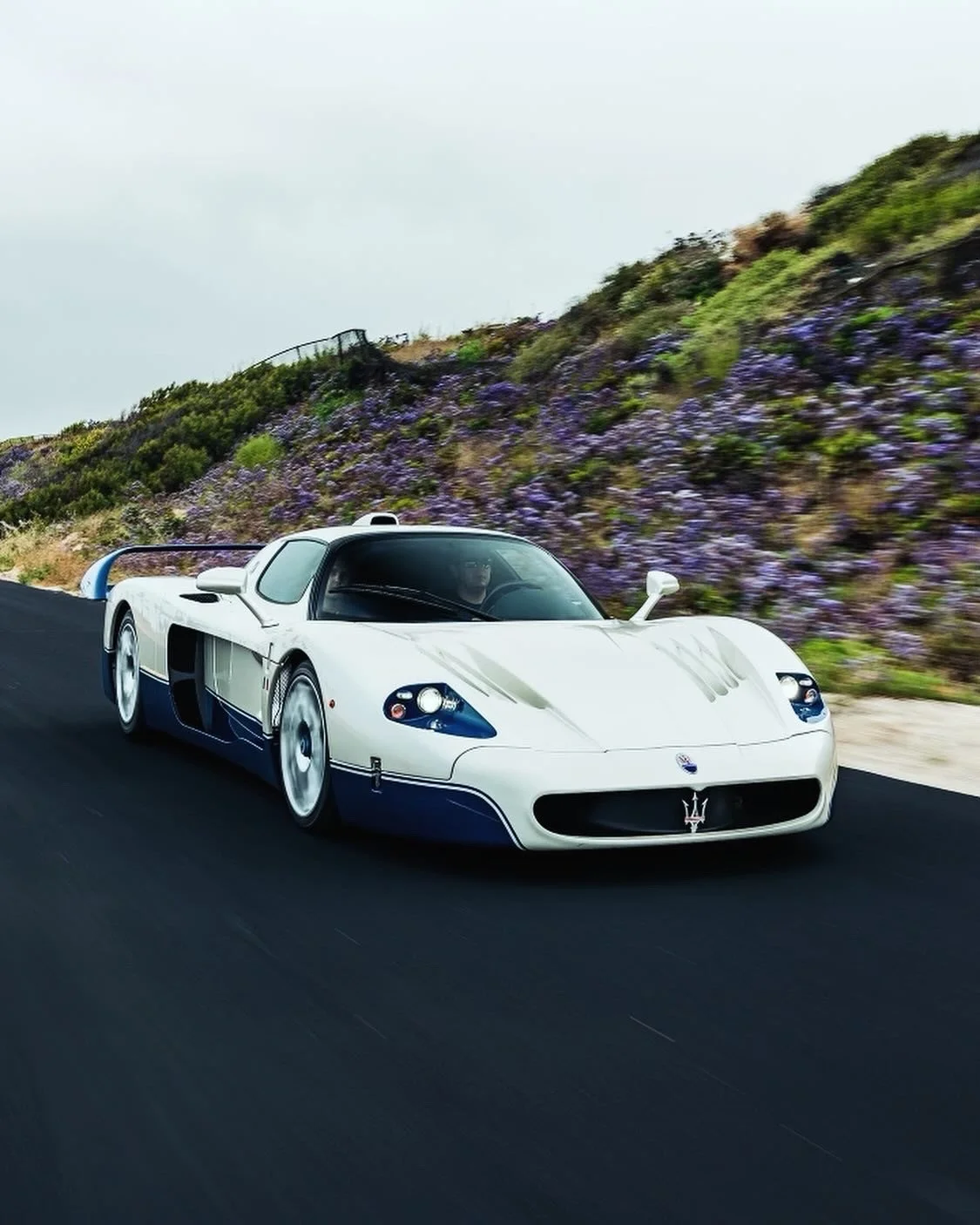 A white Maserati race car driving on a paved road beside a hillside covered with purple flowers.