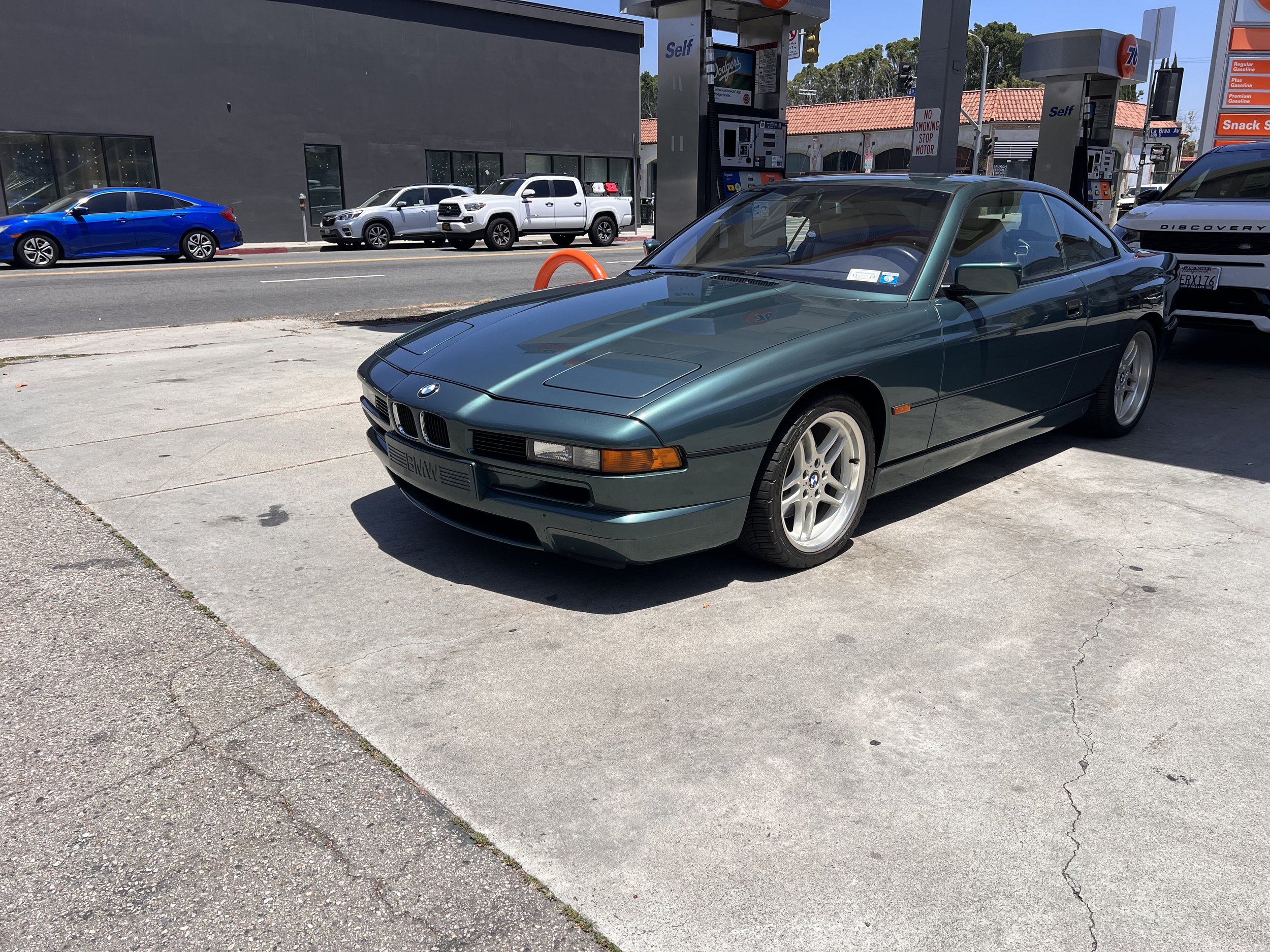 A vintage BMW car parked at a gas station with a blue and silver vehicle in the background.