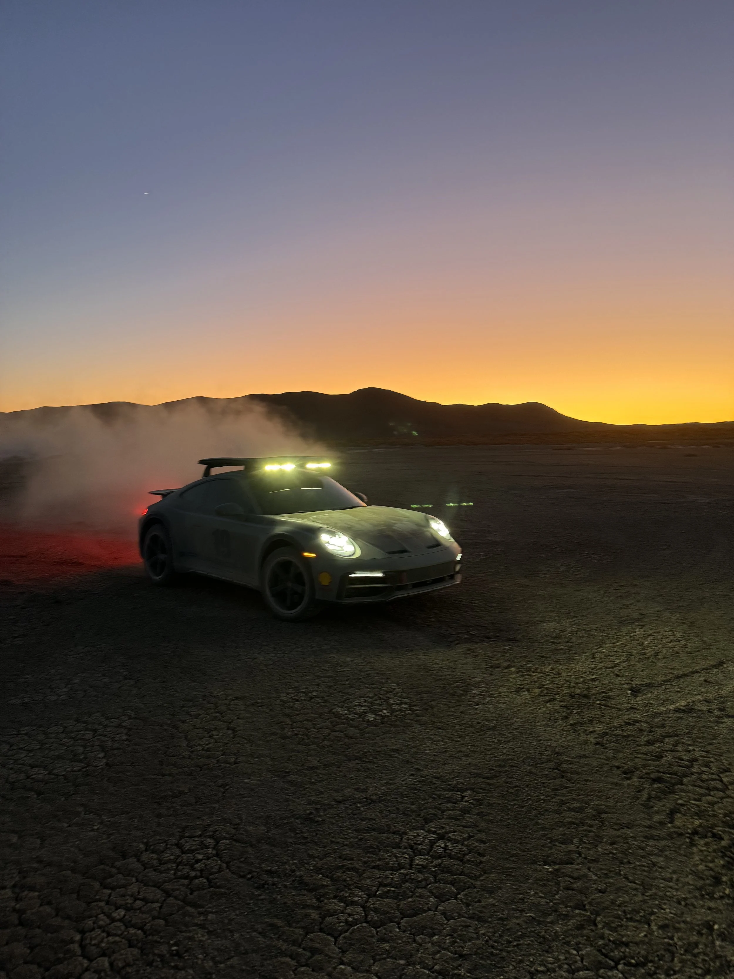 An off-road rally car with headlights and roof-mounted lights speeding on a dirt track during sunset, kicking up dust with mountains in the background.