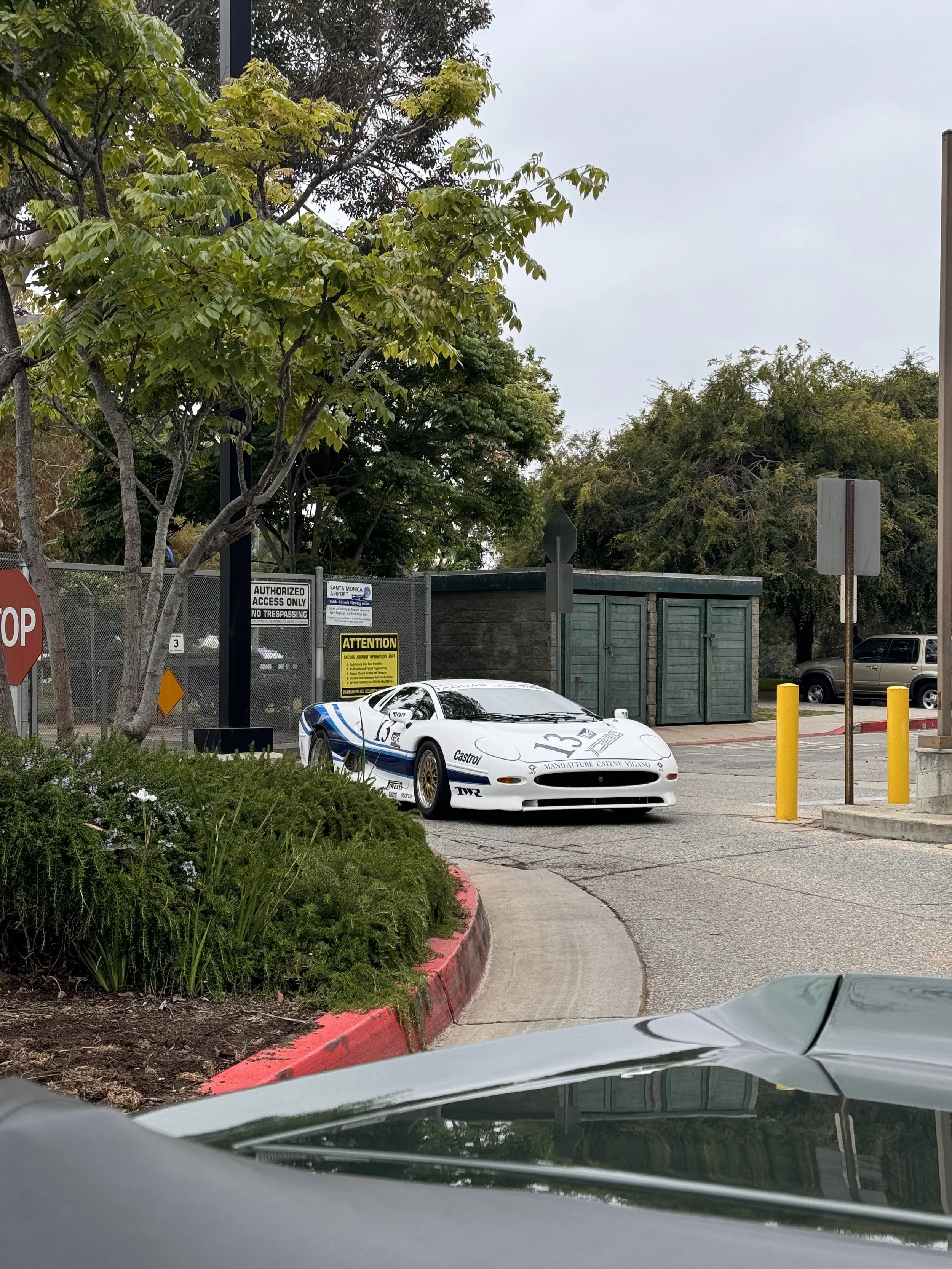 A white vintage race car with blue stripes and racing decals parked in a lot next to green trees and a small green building, with a red curb and several yellow posts around the area.
