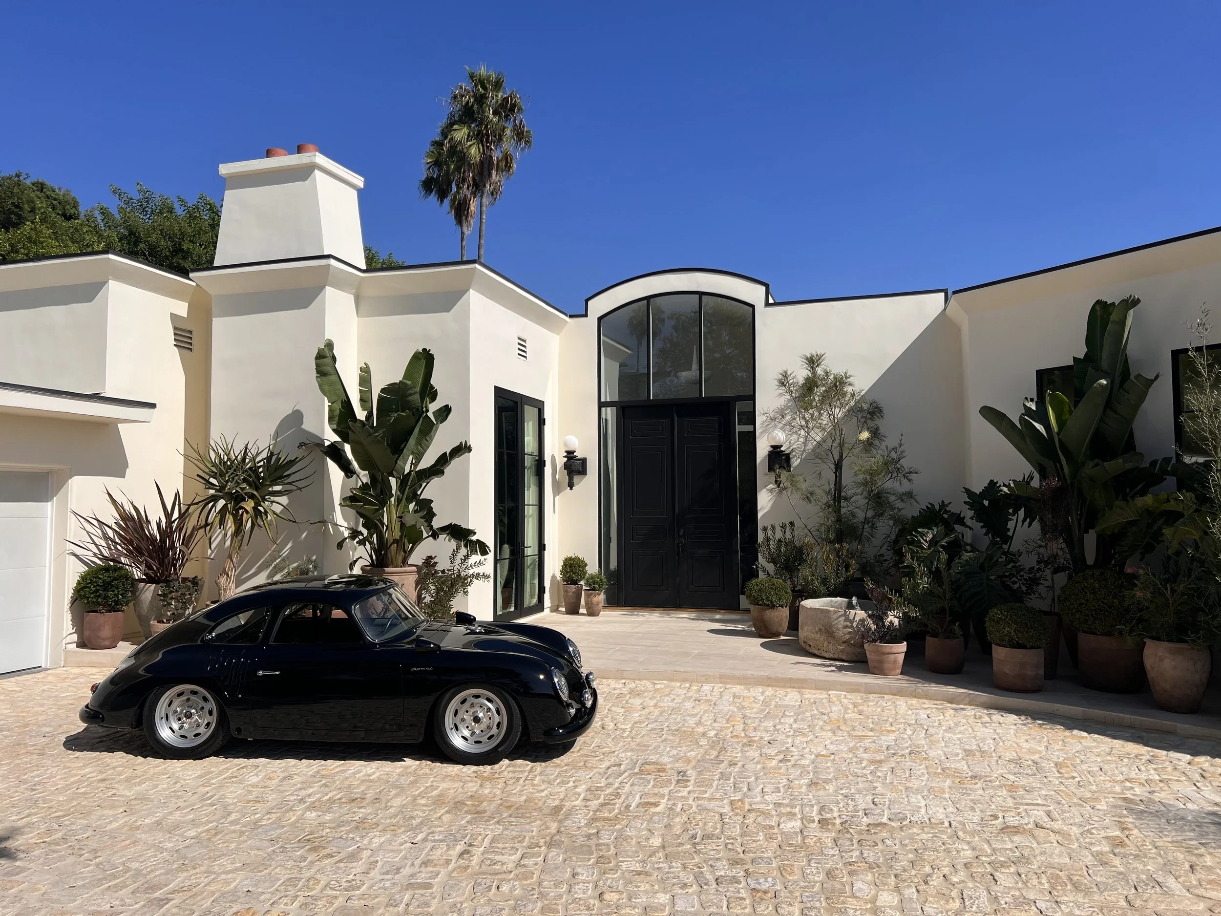 A sleek black vintage sports car parked in front of a modern white house with large black doors, potted plants, and tall palm trees under a clear blue sky.