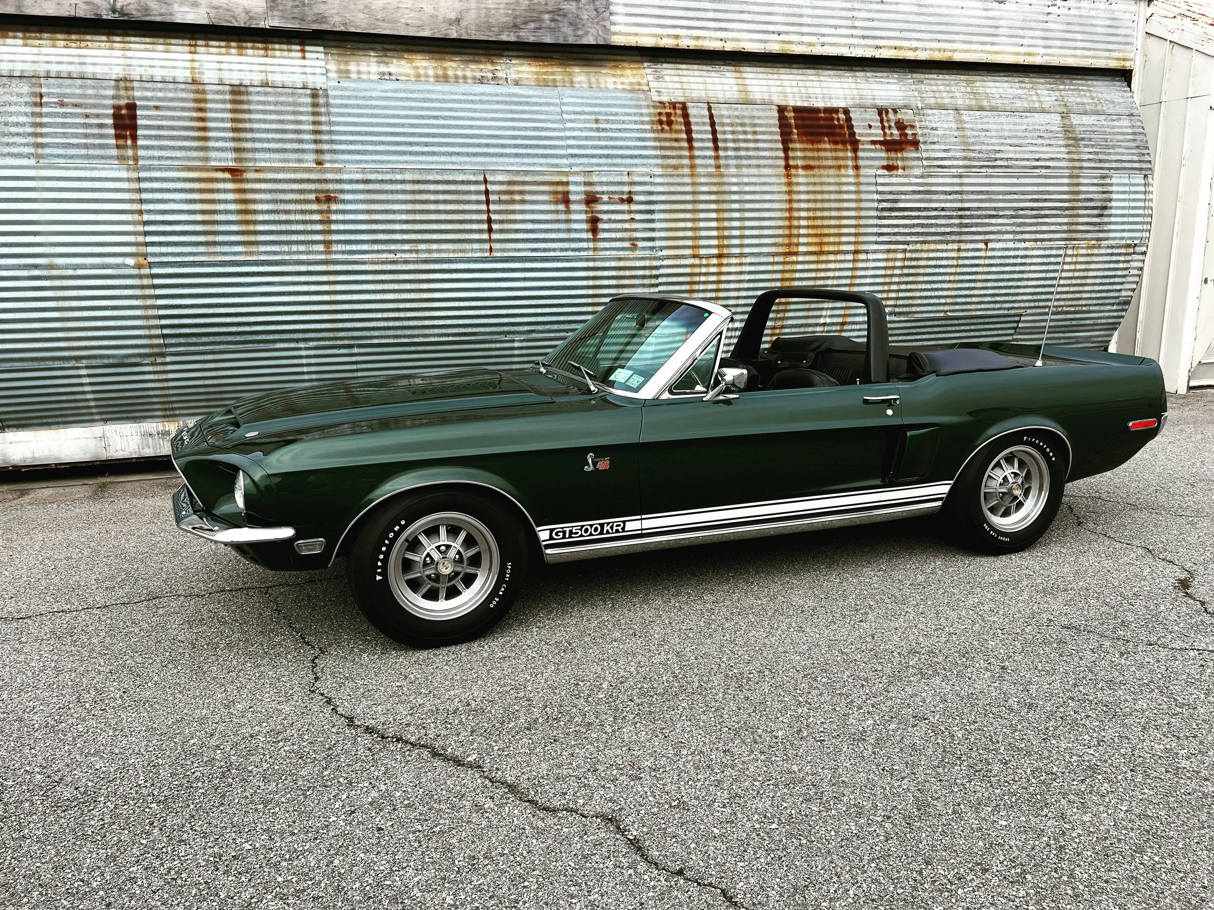 A classic dark green Ford Mustang Shelby GT500 Convertible parked on a cracked asphalt surface in front of a rusted corrugated metal wall.