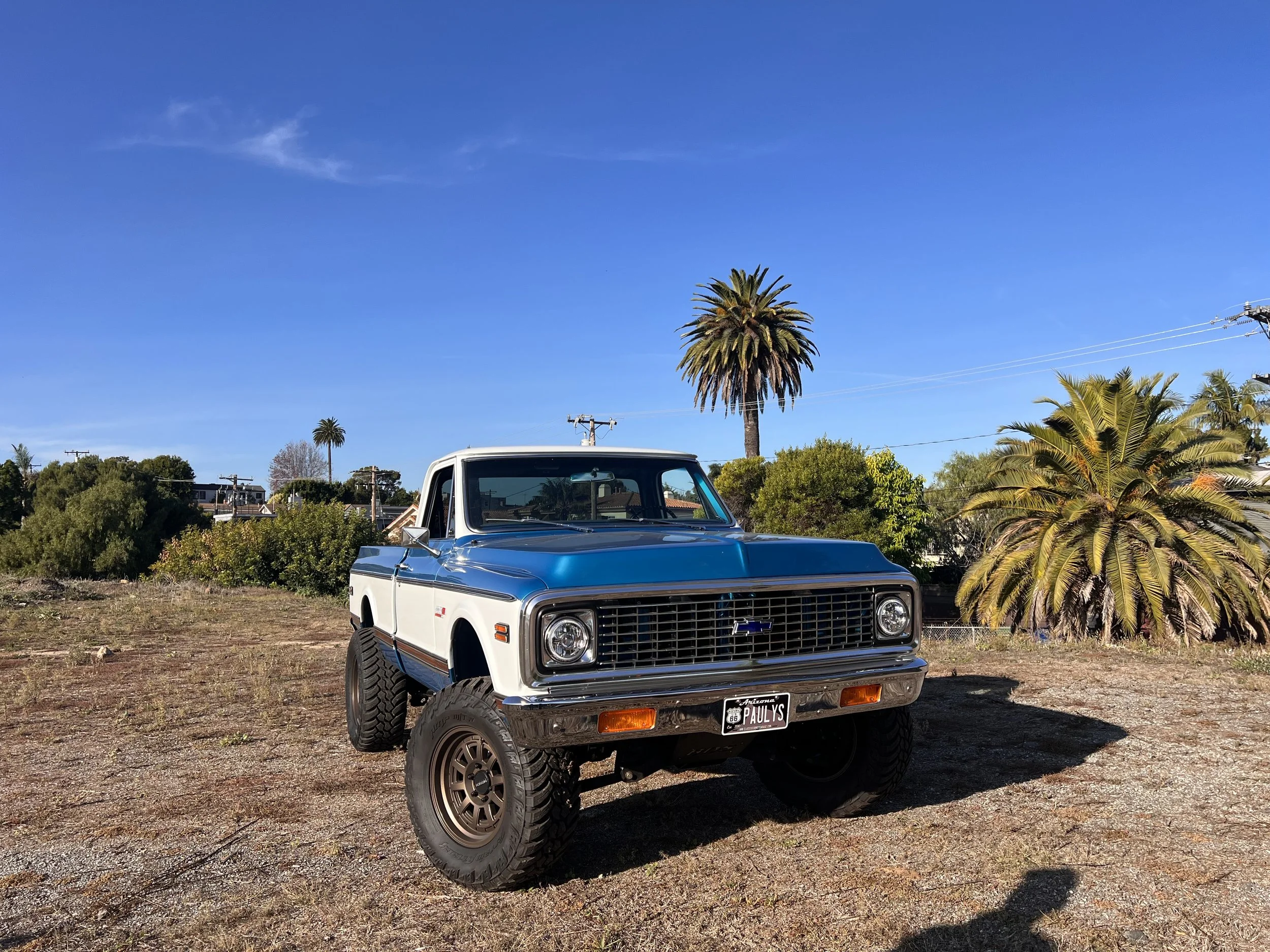 A vintage blue and white pickup truck with large tires parked on a dirt patch with green bushes and tall palm trees in the background, under a clear blue sky.