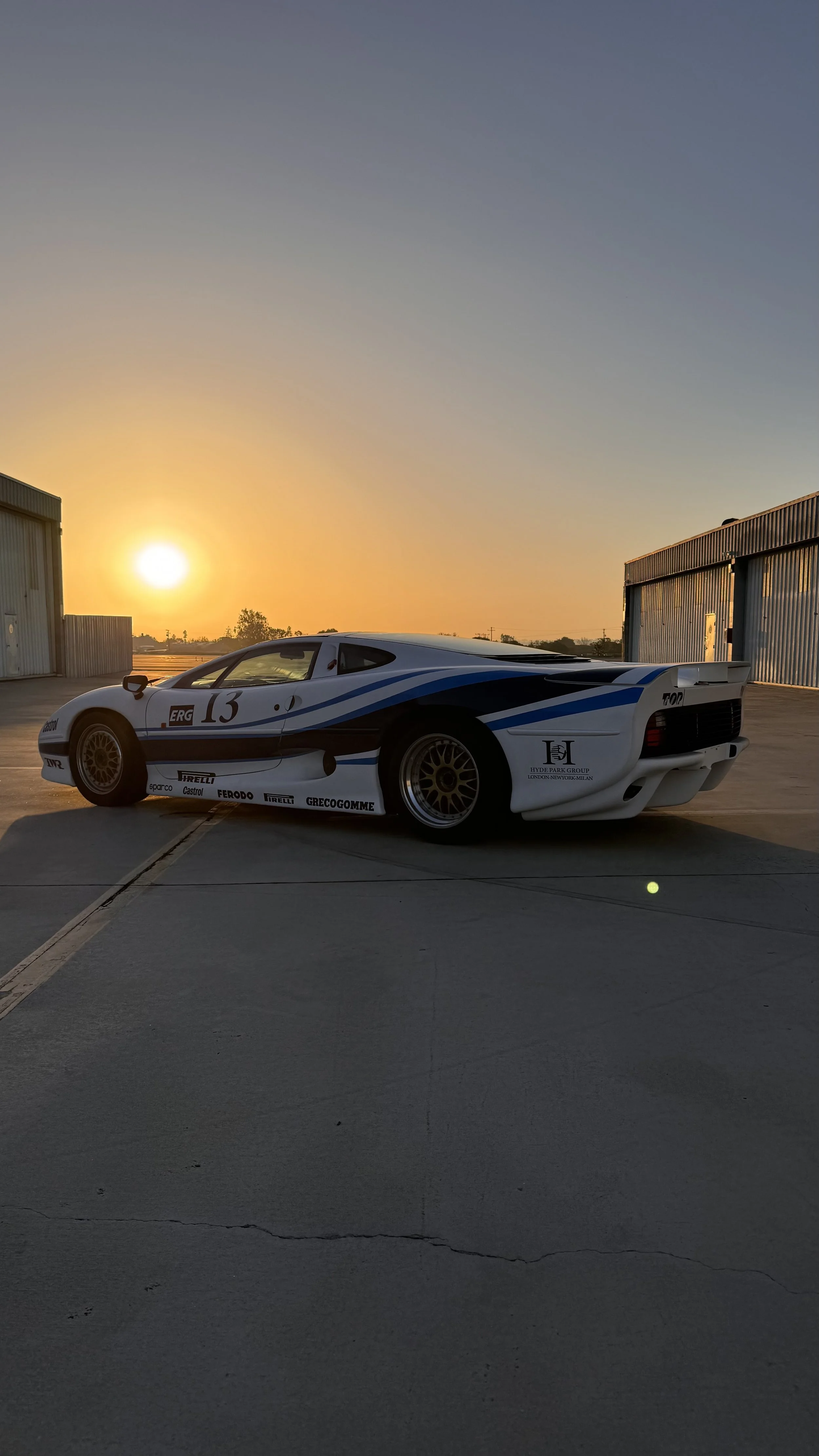 A white and blue vintage race car parked on a concrete surface at sunset, with a warehouse in the background.
