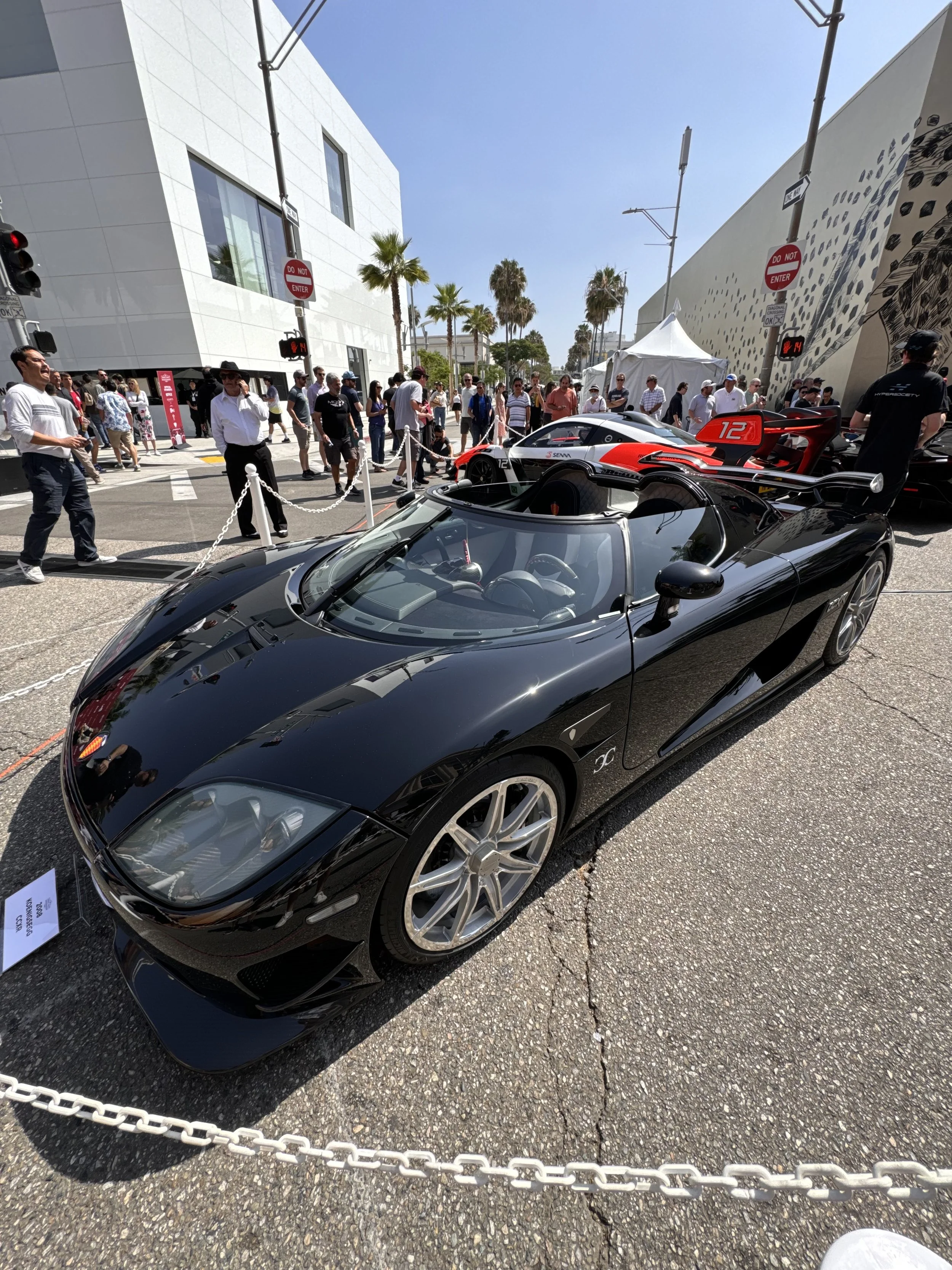 A black sports car displayed outdoors at an auto show, surrounded by people, with palm trees in the background and modern buildings.