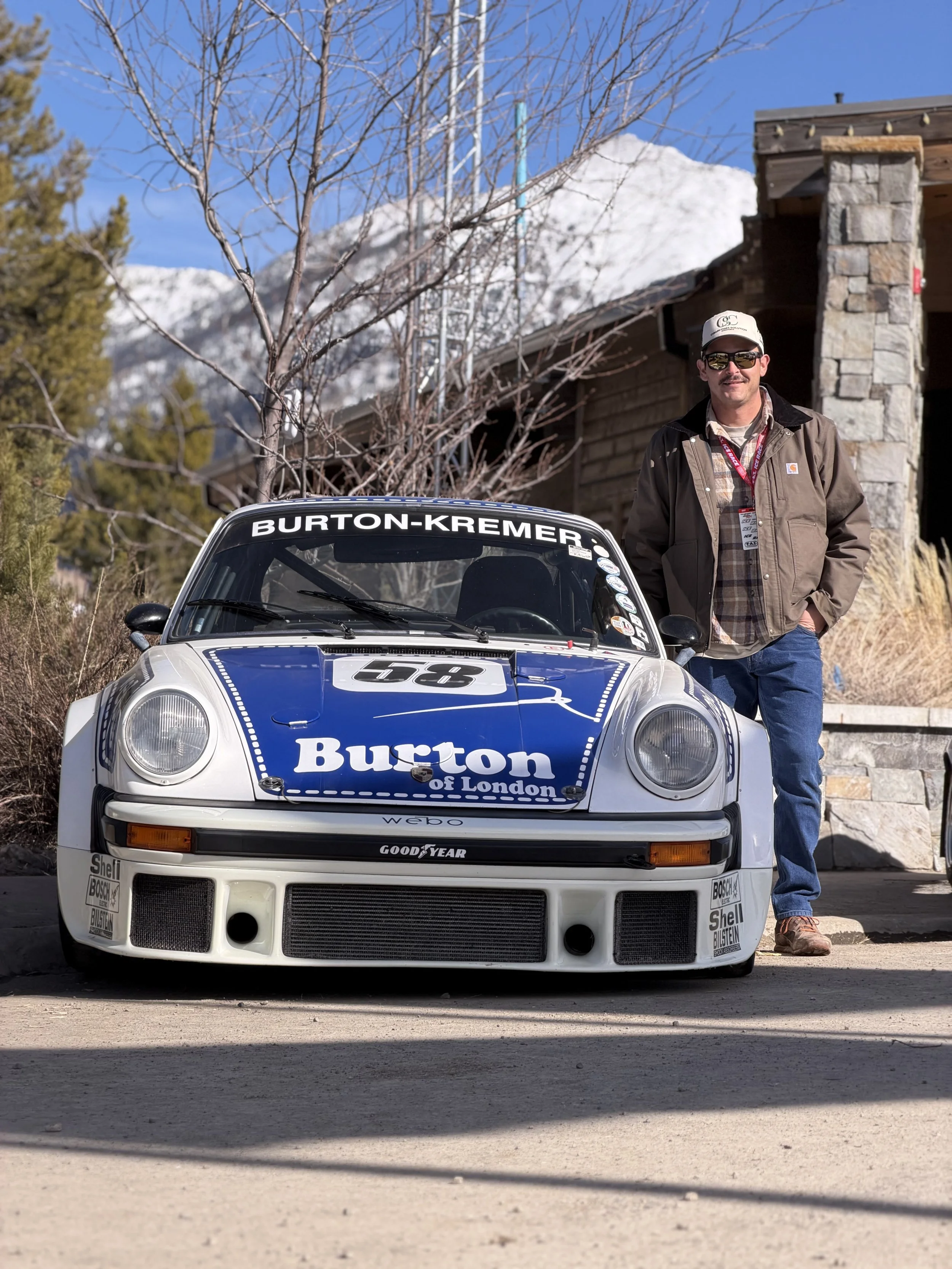 A man standing next to a vintage racing car with a mountain and a house in the background.