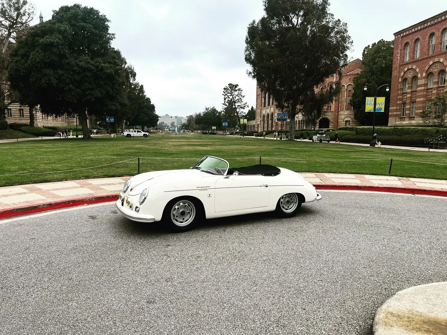 A vintage white convertible sports car parked on a street curb with green grass, trees, and brick buildings in the background.