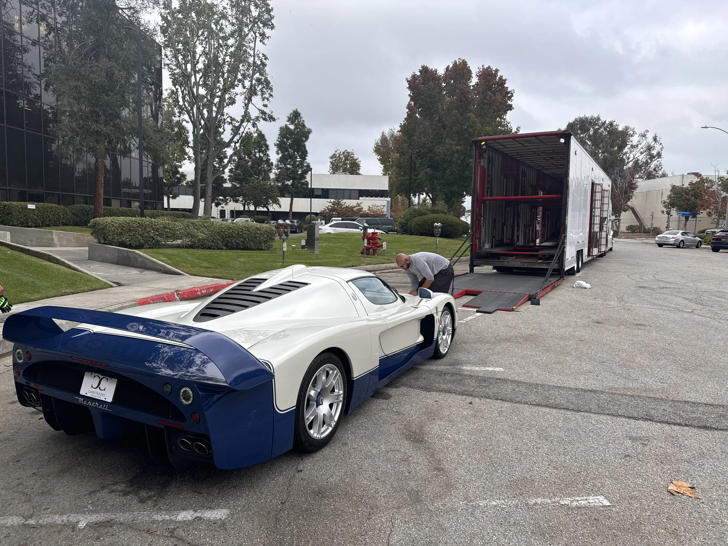 A white and blue vintage race car being unloaded from a large red transport truck with a man helping. The car is parked in an empty lot near modern office buildings and trees with changing leaves.