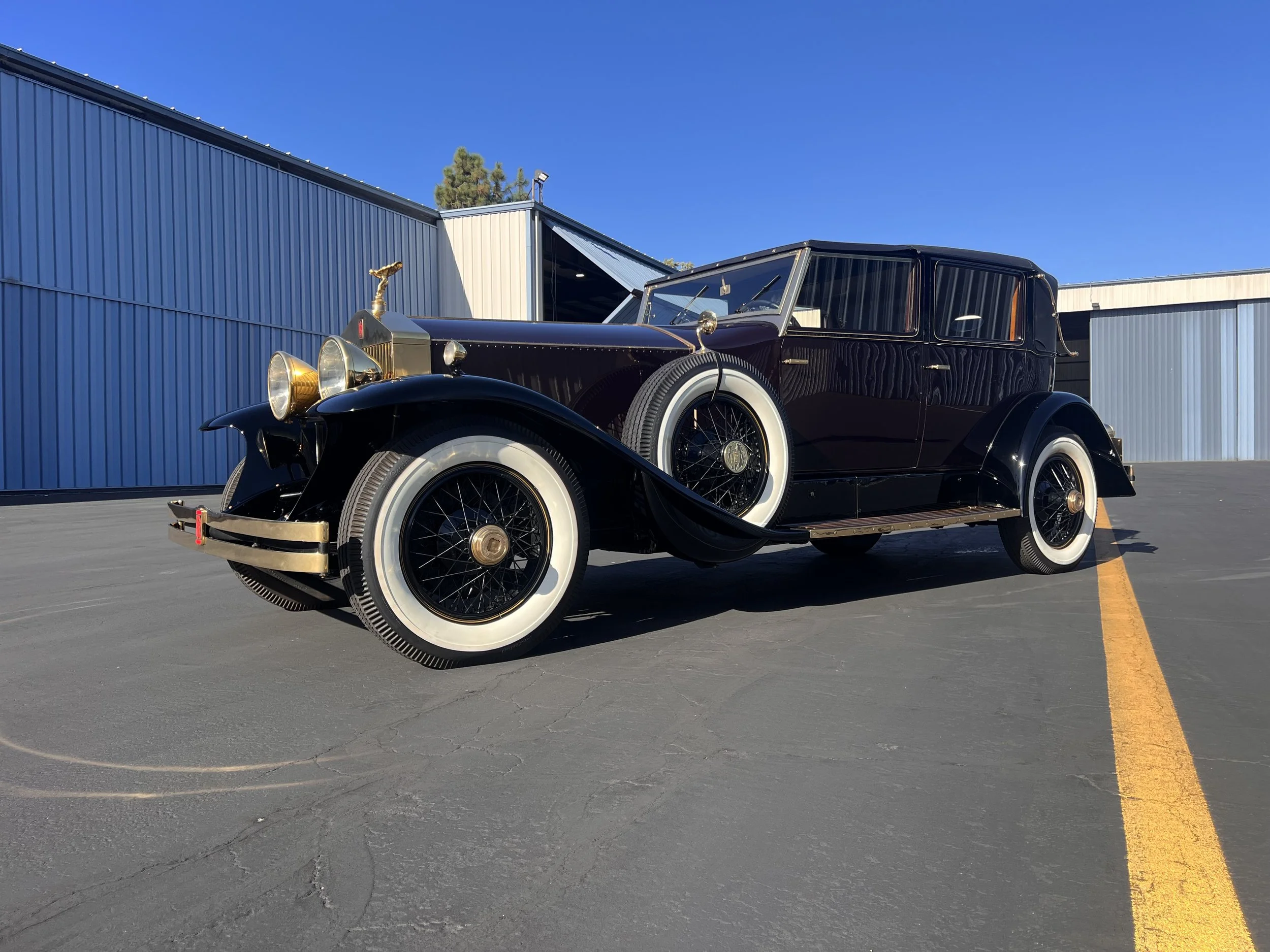 A vintage black luxury car with white tires parked on an empty asphalt lot near industrial buildings under a clear blue sky.