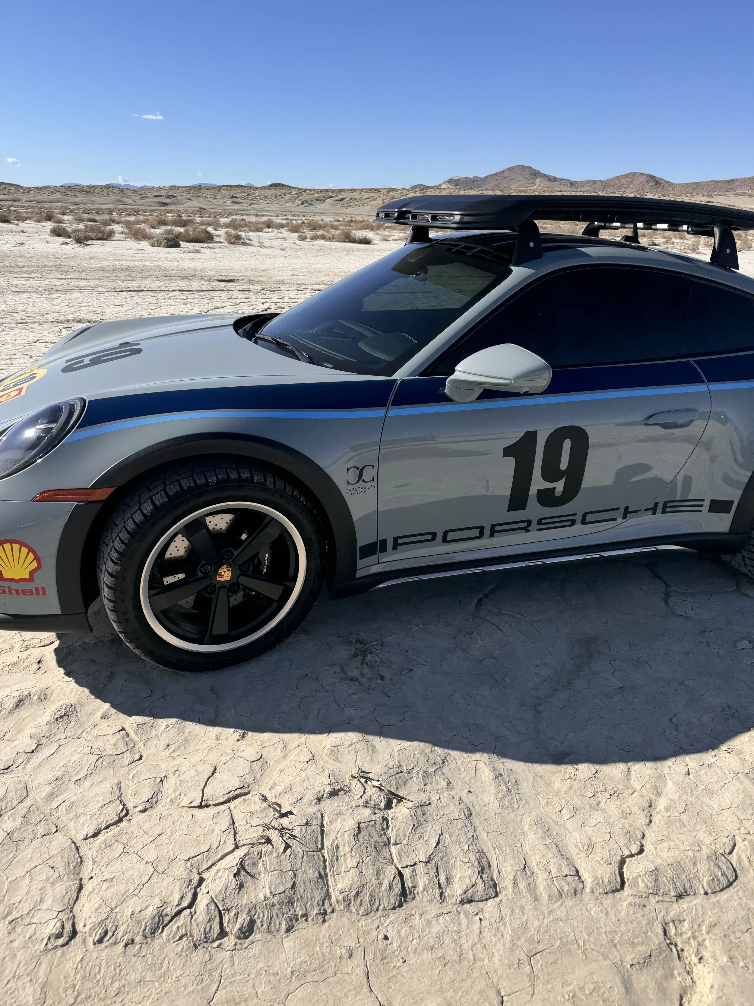 A silver Porsche race car with number 19 and sponsorship decals, parked in a desert landscape with mountains in the background.