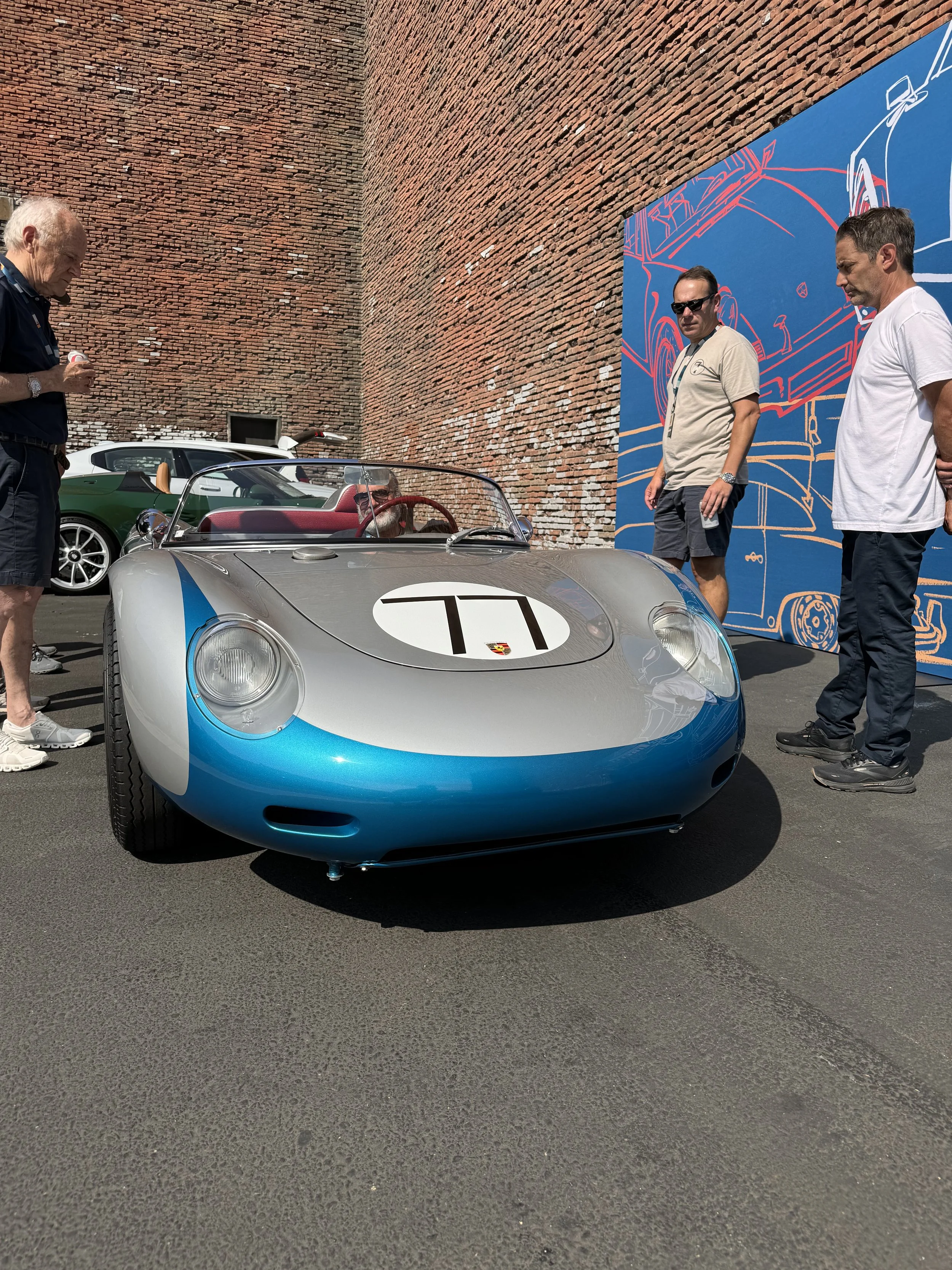 A vintage silver and blue race car with the number 71 on the hood, parked outdoors with three men looking at it, and a brick wall in the background.