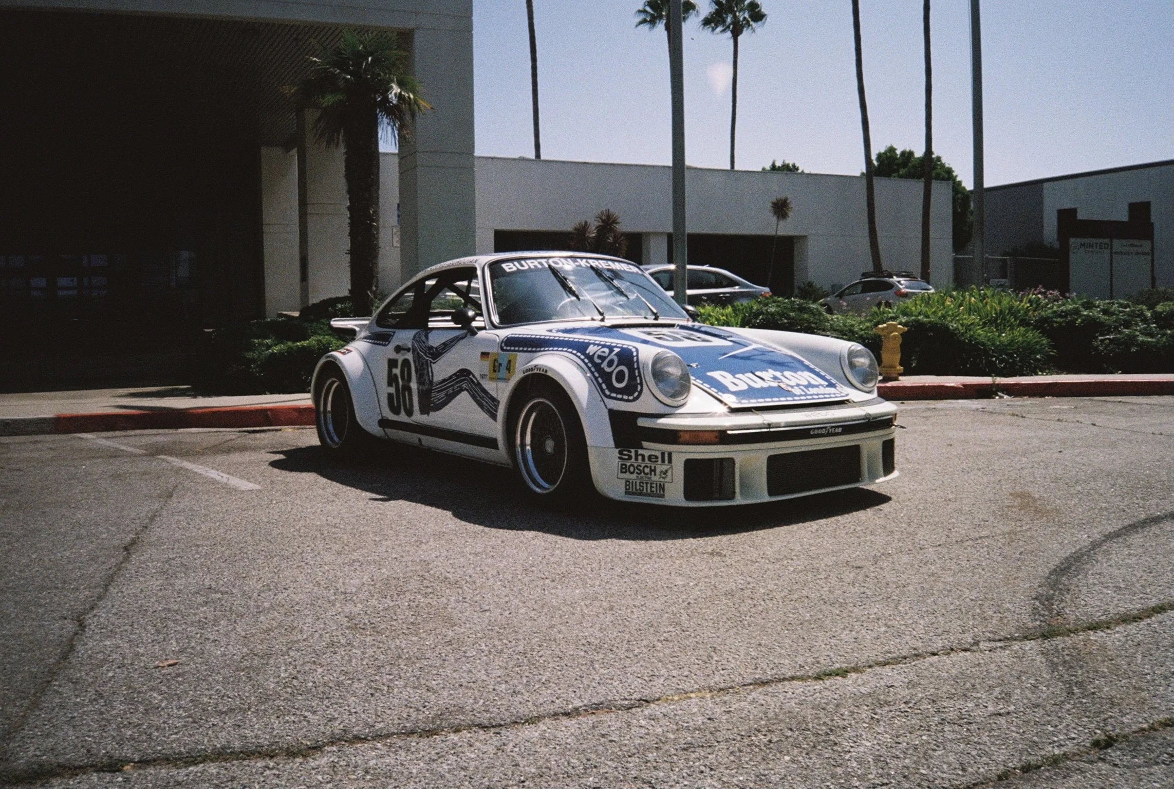 A vintage Porsche race car with the number 58, decorated with sponsor decals, parked outdoors in a lot with palm trees in the background.