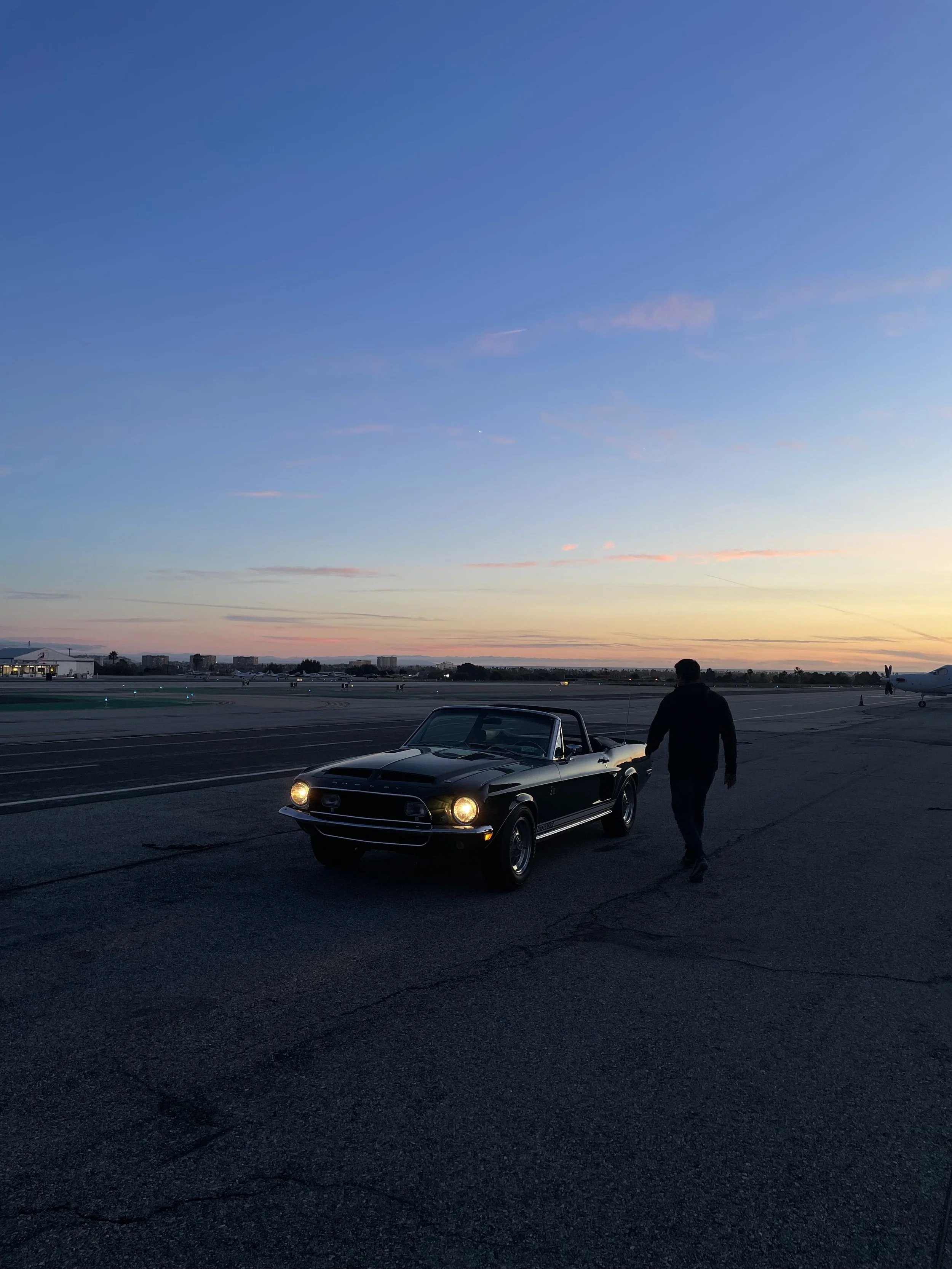 A person is walking near a black vintage Ford Mustang on the tarmac of an airport at sunset with a partly cloudy sky.