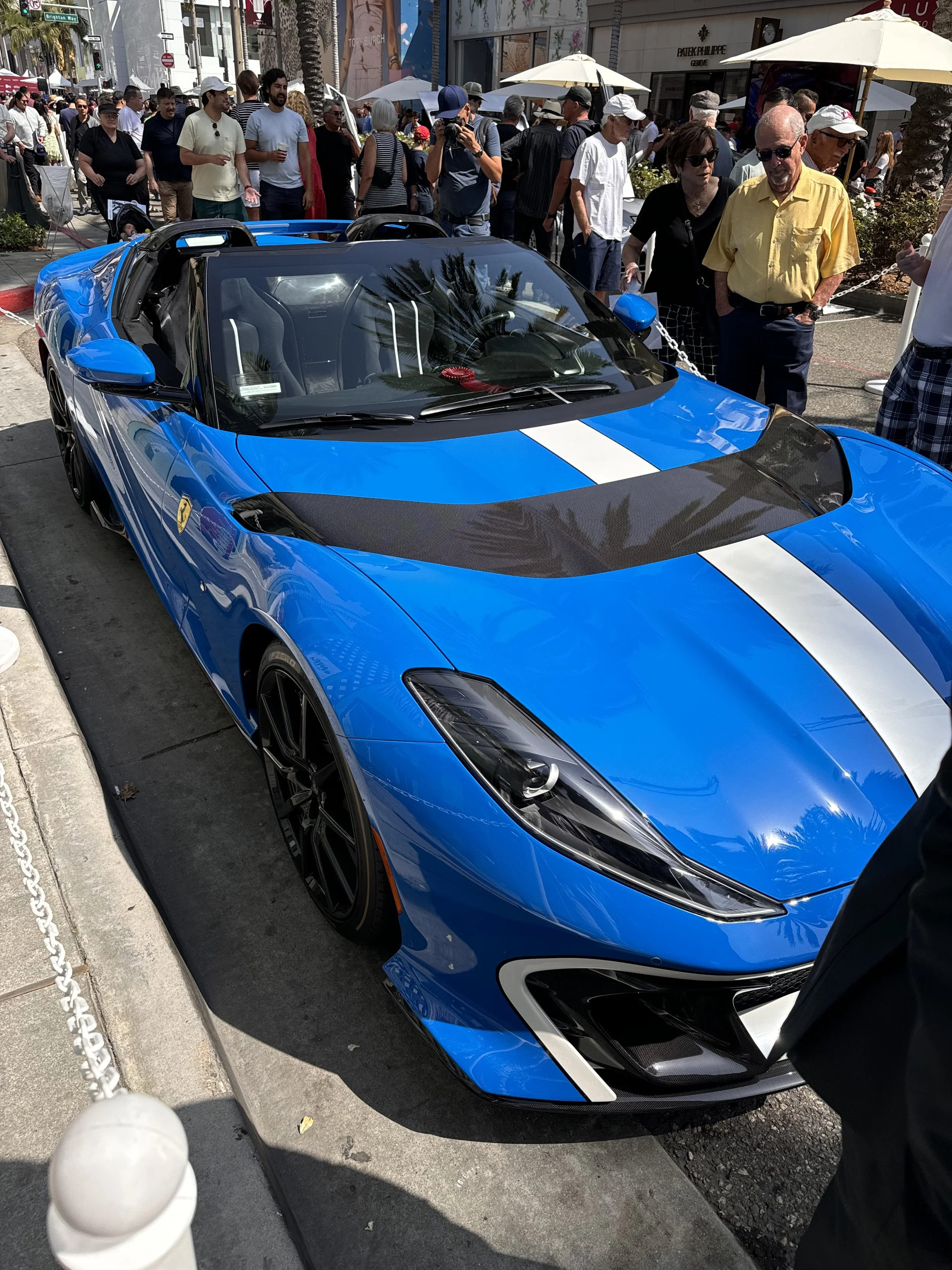 Blue sports car with a white stripe on the hood, parked on a busy sidewalk during a car show, with people gathered around.