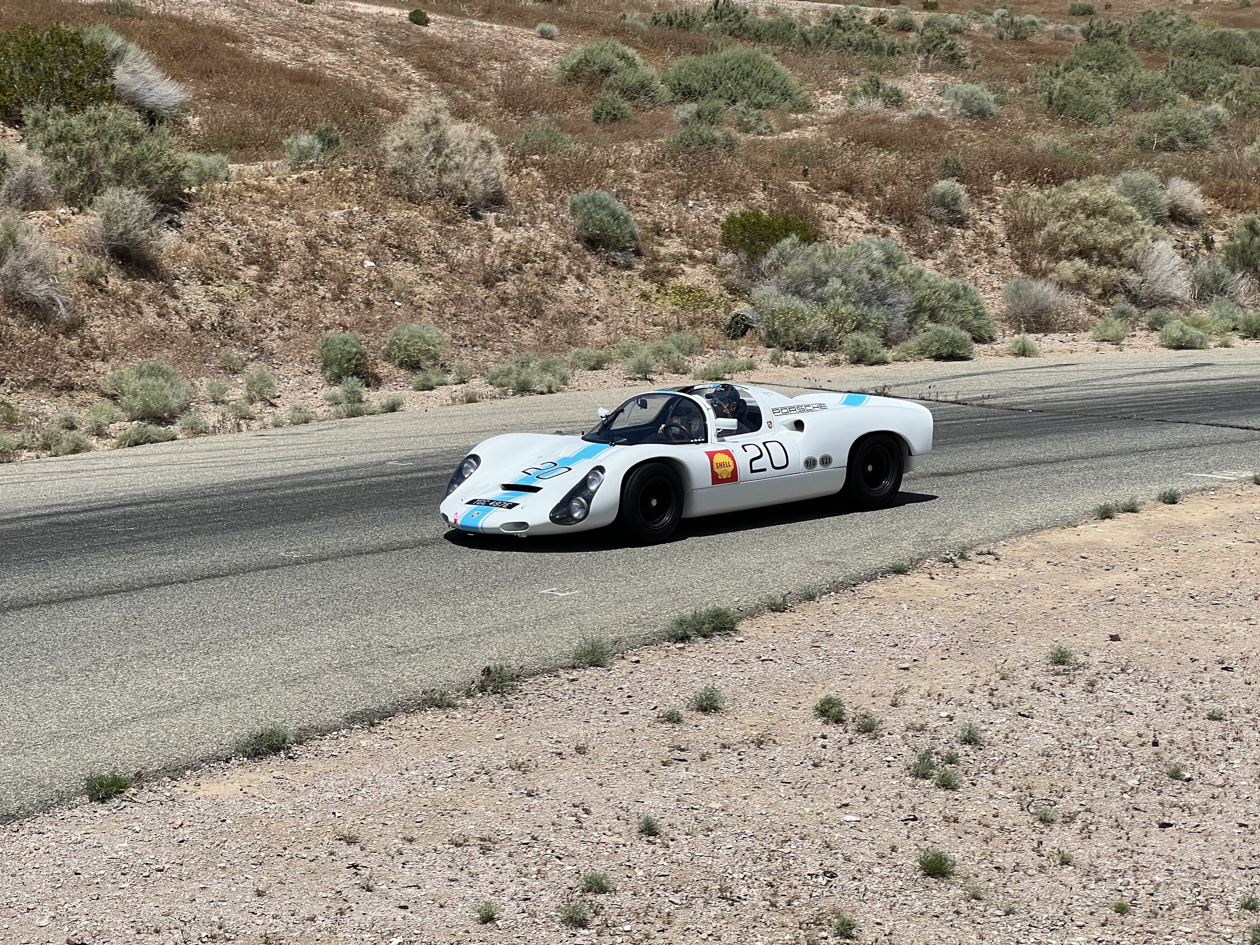 A vintage race car on a desert highway with desert landscape and low shrubs in the background.